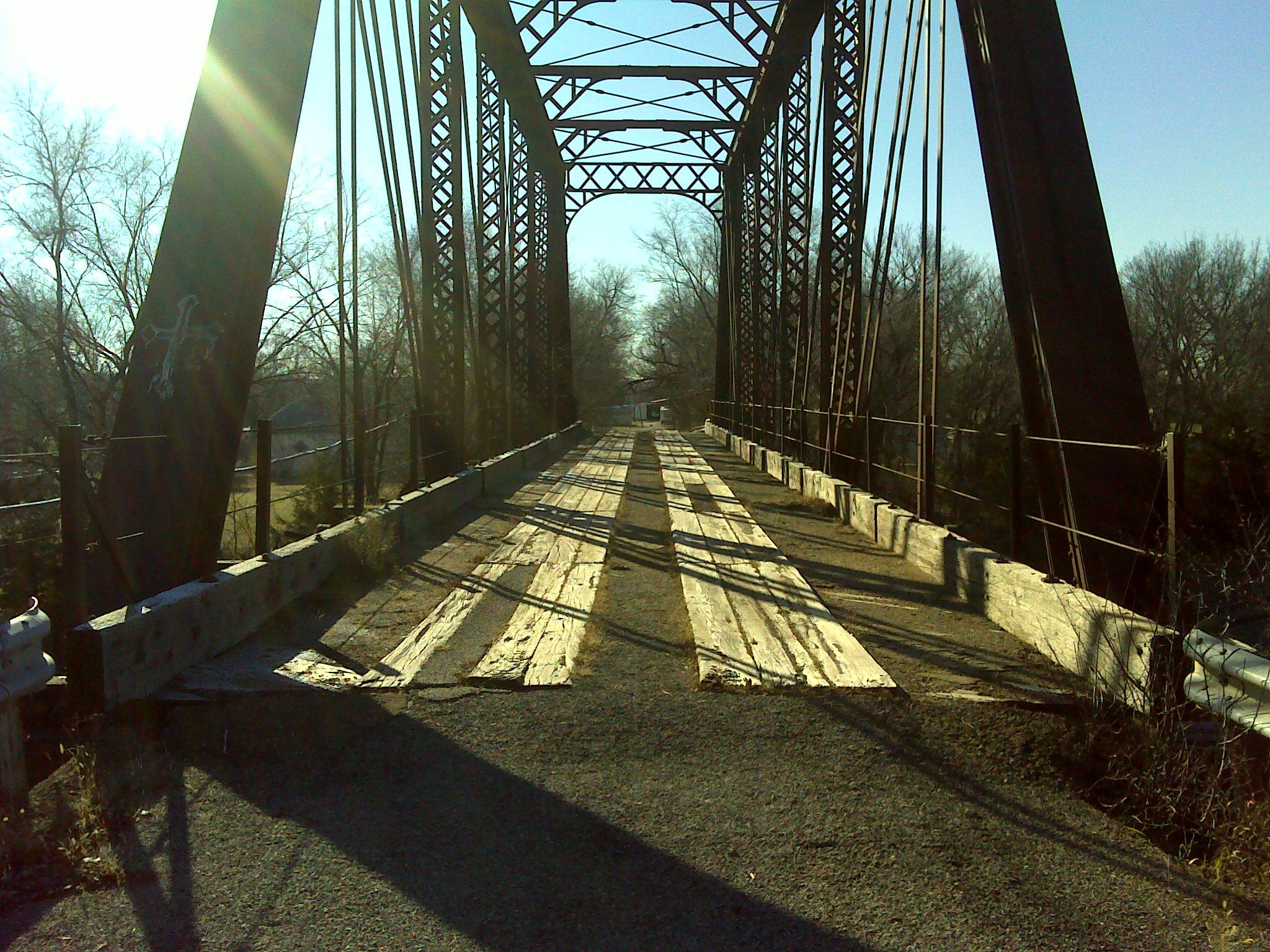 A wooden-planked bridge with a metal truss structure crossing over a dry, grassy area. The sun shines brightly in the background, casting shadows on the bridge's surface. Surrounding trees are bare, suggesting an early spring or late fall setting. Melvern Riverfront Trails mountain bike trail.
