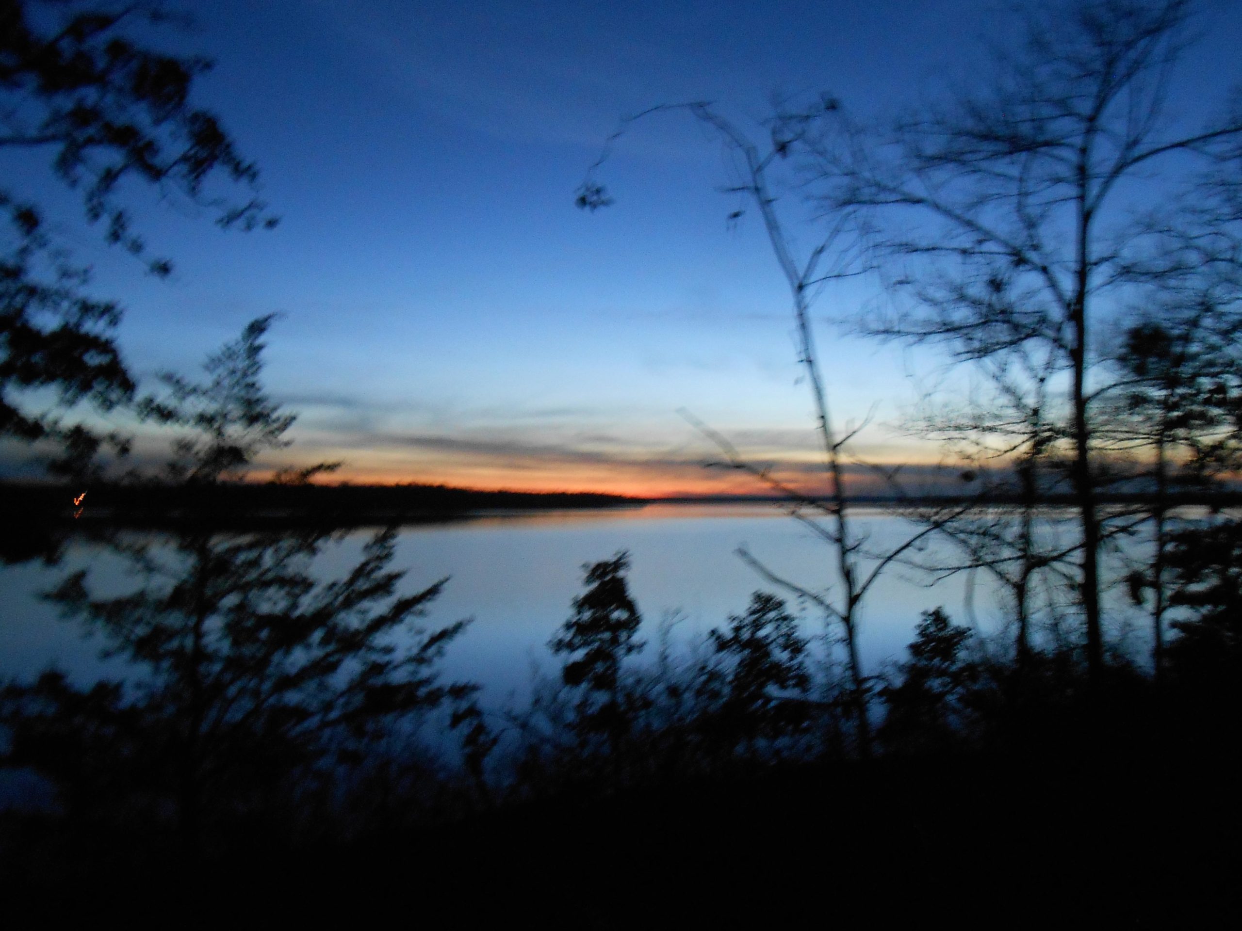 A serene twilight scene with silhouettes of trees framing a calm body of water reflecting shades of blue and orange in the sky. The horizon hints at the transition between day and night. Barber Hills Trail at Pat Mayse Lake mountain bike trail.