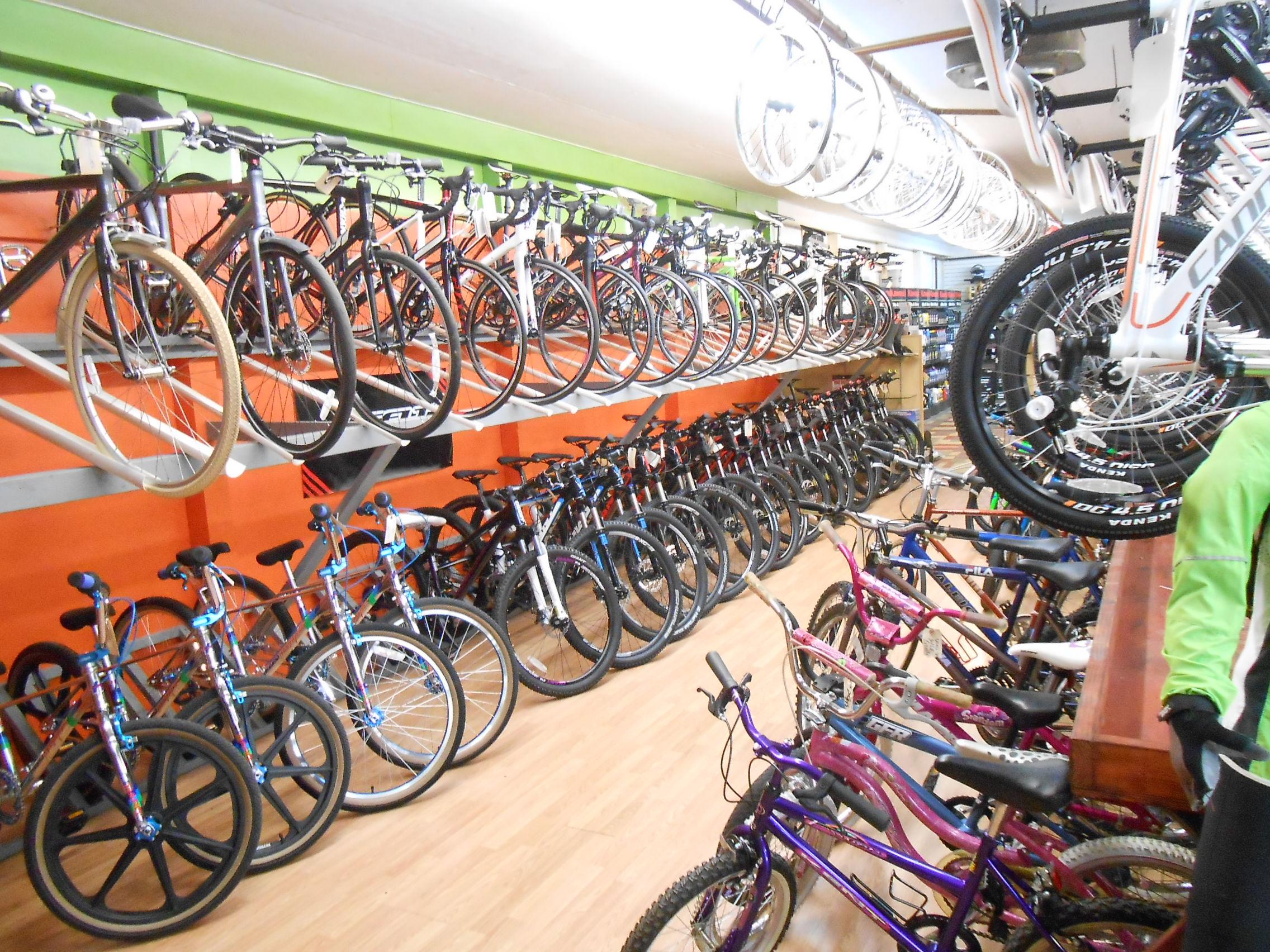 A view of a brightly colored bike shop showcasing a variety of bicycles. The shop features multiple rows of bikes, including road bikes and mountain bikes, displayed on racks above and on the floor. Large wheels are hanging from the ceiling, adding to the vibrant atmosphere of the store.