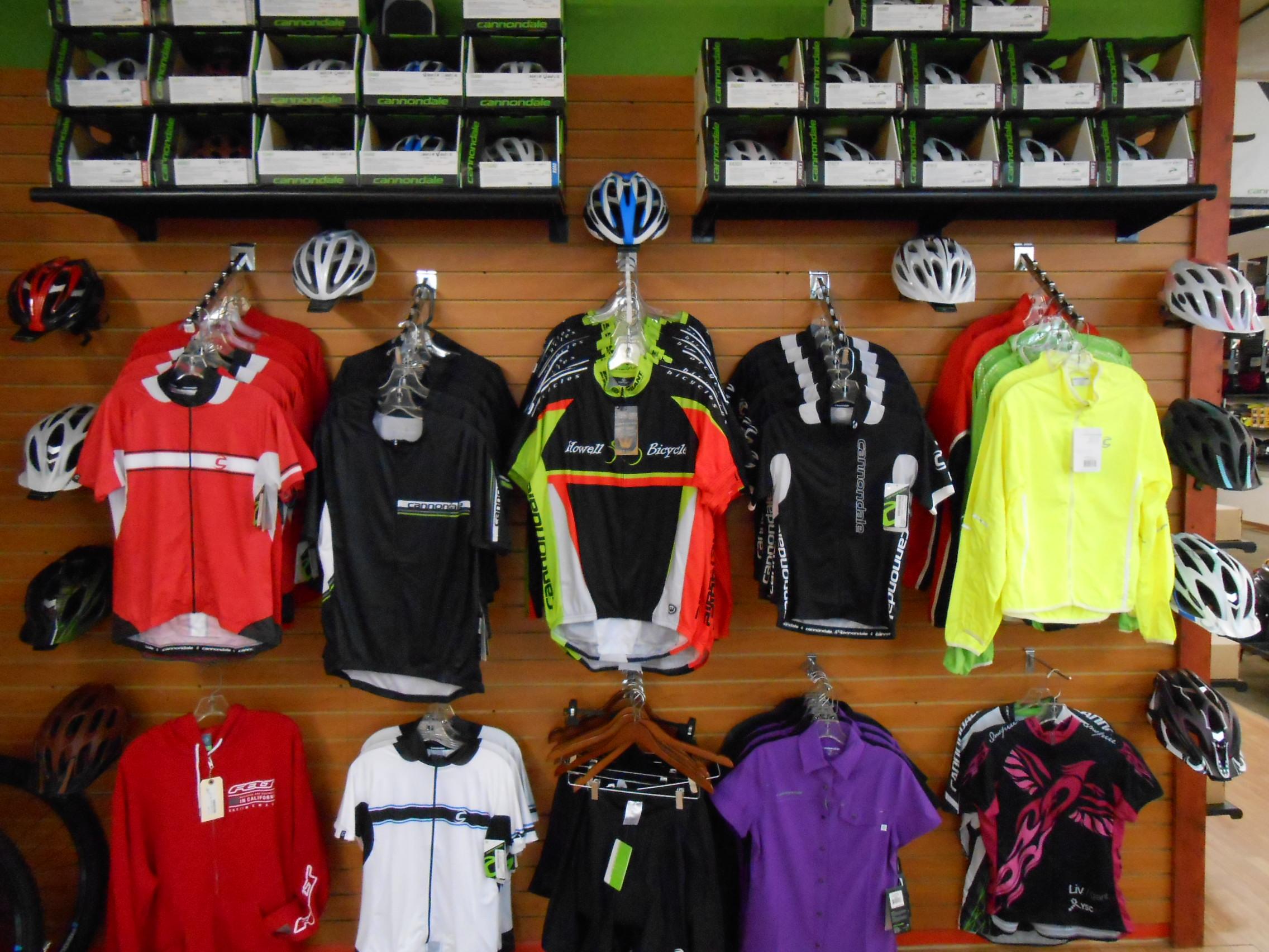 A display of cycling apparel and helmets in a bike shop. The wall features various jerseys in red, black, green, and yellow, along with a selection of helmets mounted above. Several cycling shirts hang from hooks, while more cycling gear is visible on shelves above. The store's interior is brightly lit, emphasizing the vibrant colors of the clothing.