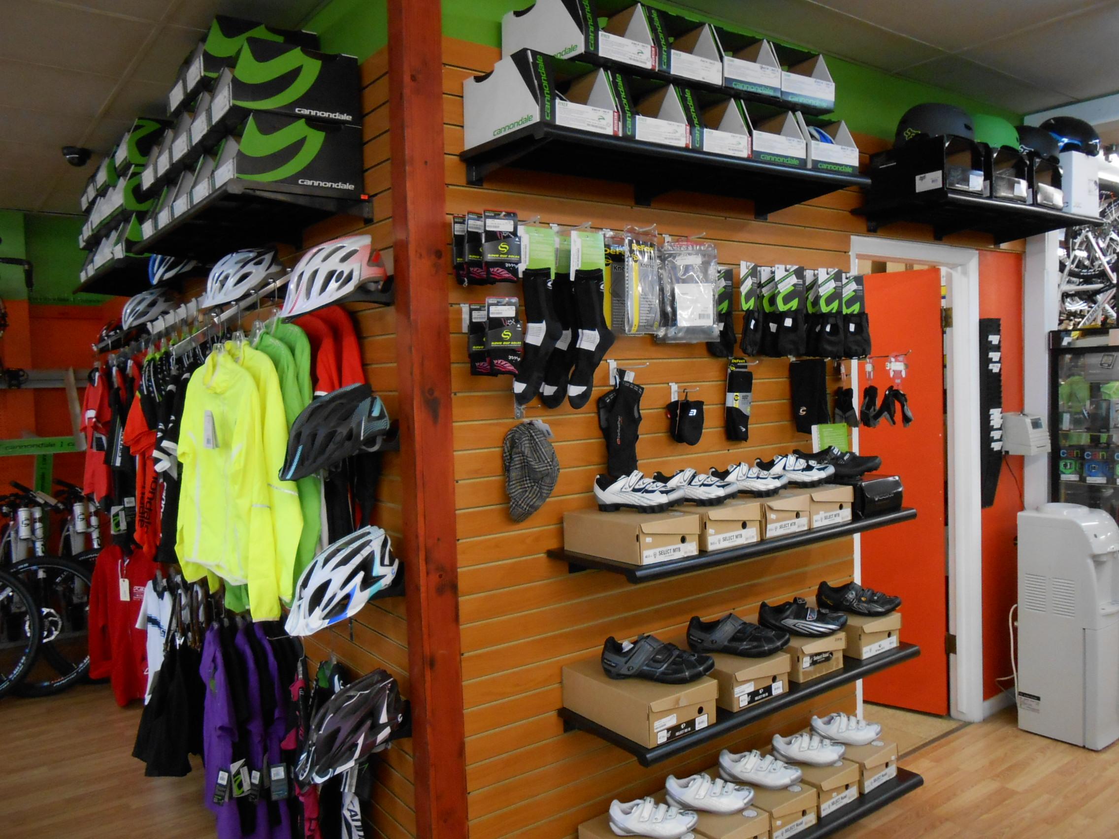 A well-organized display in a bike shop featuring a variety of cycling gear. The wall showcases brightly colored jackets, helmets, and cycling shoes neatly arranged on shelves. Accessories such as socks and gloves are also visible alongside helmets. The shop's interior is bright and inviting, with an orange door and wooden paneling in the background.