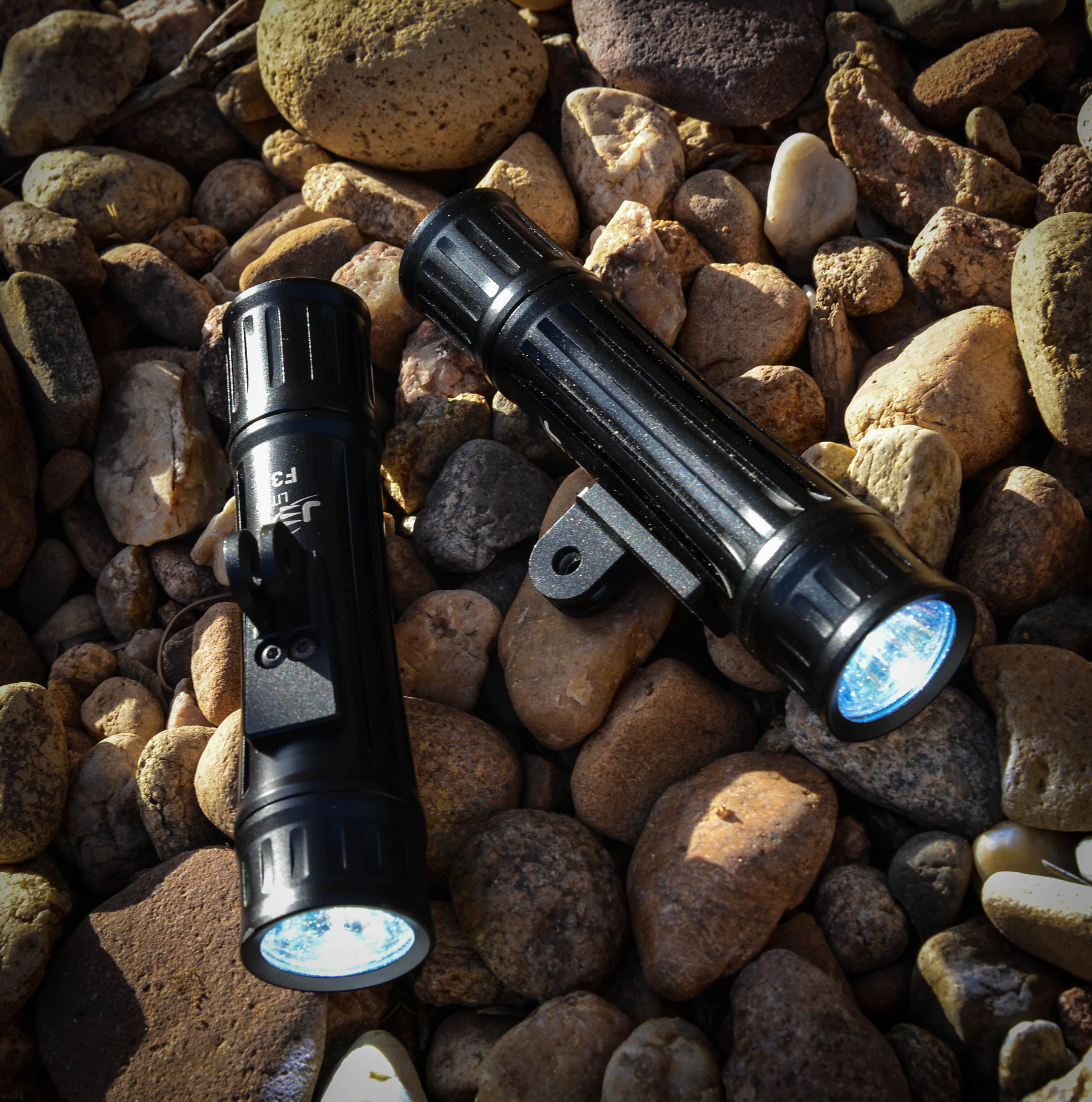Two black flashlights are positioned on a bed of smooth pebbles, with one flashlight angled slightly to show its front. The flashlights feature textured grips and bright LED lights that are illuminated. The surrounding environment is natural, characterized by various sizes and colors of rocks.