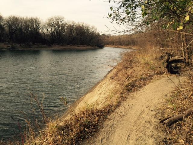 A serene riverbank scene featuring a calm river winding through a sandy shore. Bare trees are visible along the banks, creating a tranquil natural environment. The landscape is framed by sparse vegetation, suggesting a late autumn or early winter setting. Bloomington Ferry Trail mountain bike trail.
