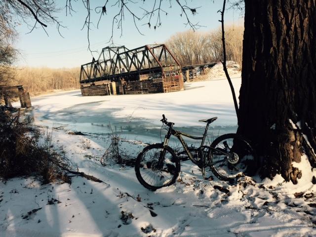 Bicycle leaning against a tree near a frozen river, with an abandoned bridge in the background. The scene is set in a winter landscape, featuring snow-covered ground and bare trees. Bloomington Ferry Trail mountain bike trail.