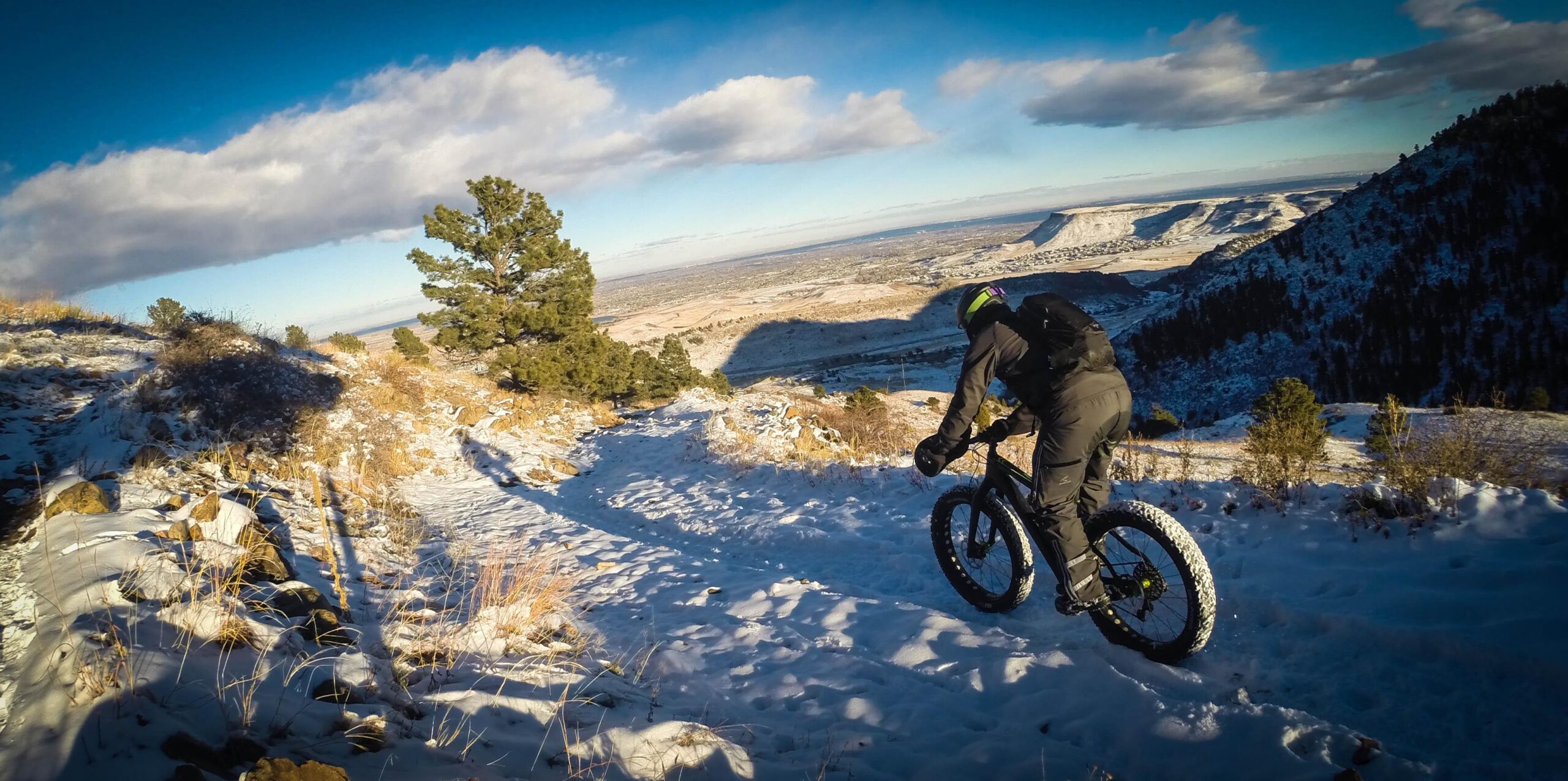 A cyclist riding a fat bike on a snowy mountain trail, with a scenic view of valleys and hills in the background under a clear blue sky. The rider is dressed in warm, dark clothing and a helmet, navigating through the snow-covered terrain amidst patches of grass and scattered rocks. White Ranch mountain bike trail.