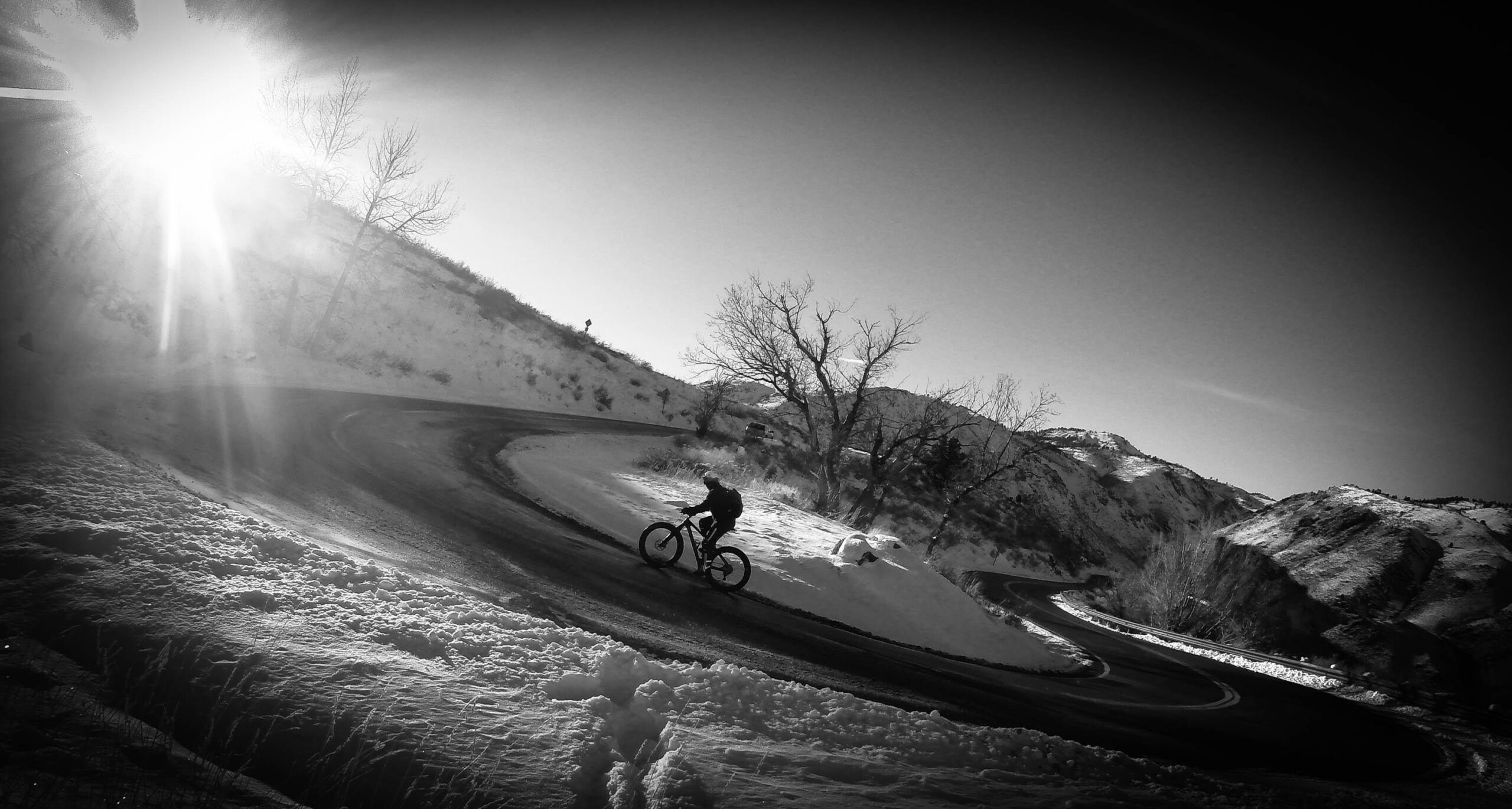 A cyclist riding a mountain bike along a winding road surrounded by snow-covered hills, with the sun shining brightly in the background. The scene is captured in black and white, highlighting the curves of the road and the silhouette of the cyclist against the winter landscape. Chimney Gulch mountain bike trail.