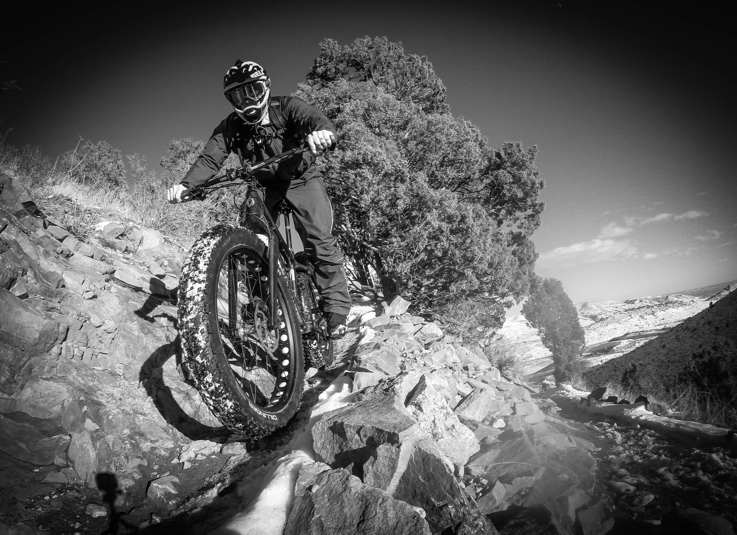 A mountain biker navigating a rocky trail surrounded by greenery, captured in black and white. The biker, wearing a helmet and goggles, is riding a fat bike with wide tires, moving along a rugged path that includes loose stones and a dusting of snow. The background features a scenic landscape with rolling hills and a partially cloudy sky. Apex Park mountain bike trail.