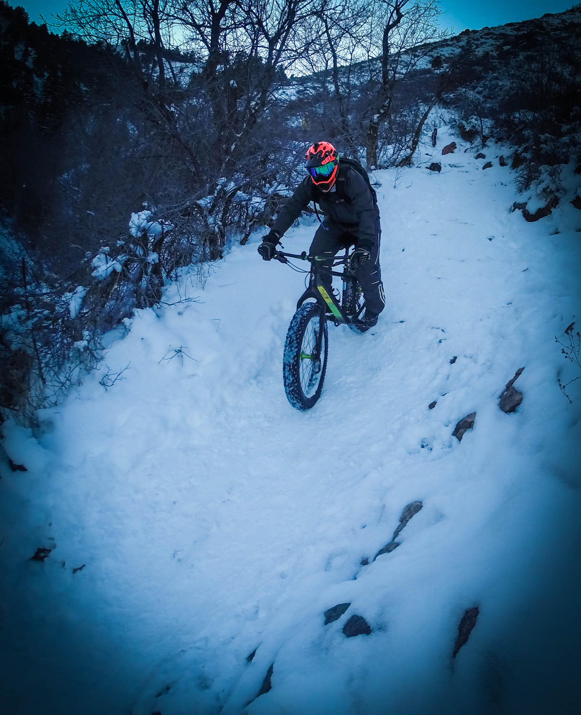 A person riding a fat tire mountain bike down a snowy trail surrounded by bare trees and rocky terrain. The cyclist is wearing a helmet and goggles, dressed in winter gear, as they navigate the snowy path. The scene is set in a mountainous area with a chilly, overcast atmosphere. Apex Park mountain bike trail.