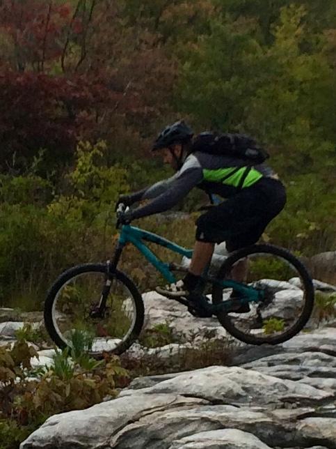 A mountain biker navigating a rocky trail surrounded by greenery and autumn foliage. The rider is wearing a helmet and a black and green outfit, focused on maintaining balance while biking on uneven terrain. CVI Trails mountain bike trail.
