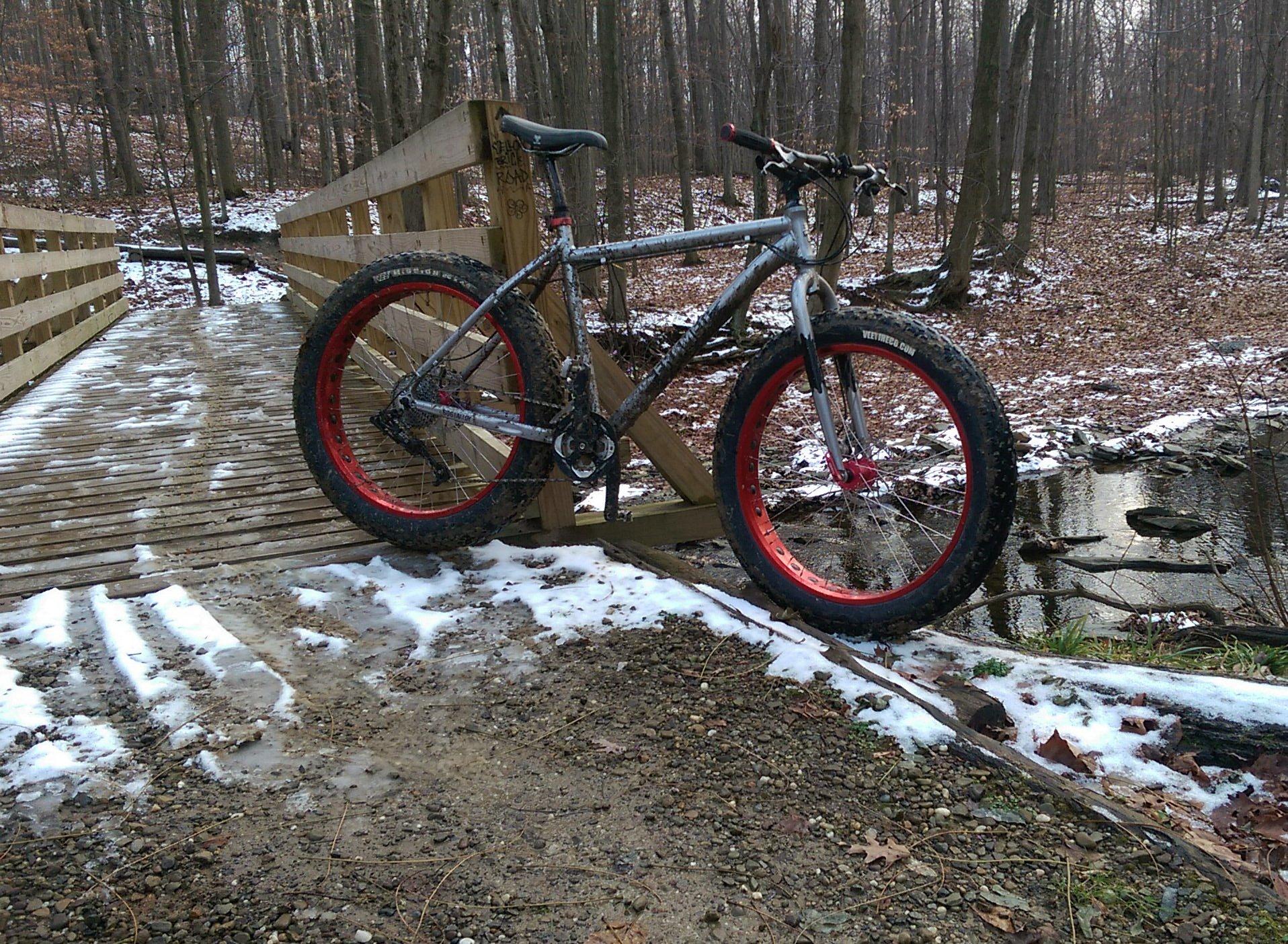 Framed Framed 2.0: A fat bike with red rims is resting on a wooden bridge in a forested area, surrounded by fallen leaves and patches of snow. A small creek is visible to the right of the bridge.