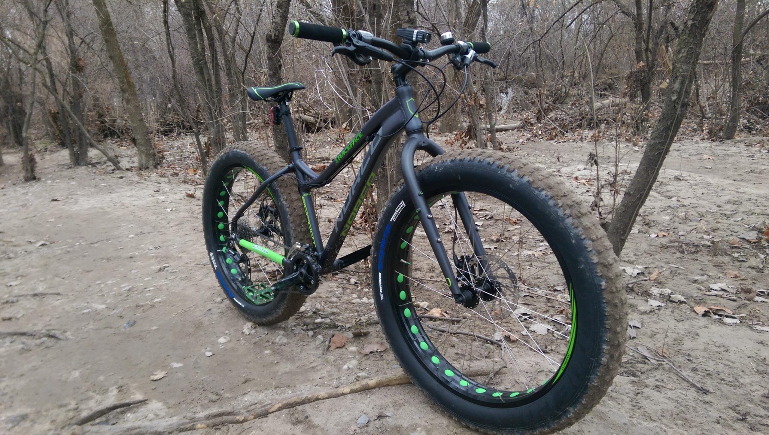 Norco Bigfoot: A black and green fat bike is positioned on a dirt path surrounded by trees. The bike features oversized tires with green accents, rugged treads, and a sturdy frame designed for off-road riding. The scene captures a natural setting with bare branches and scattered leaves, indicating a winter or early spring environment.