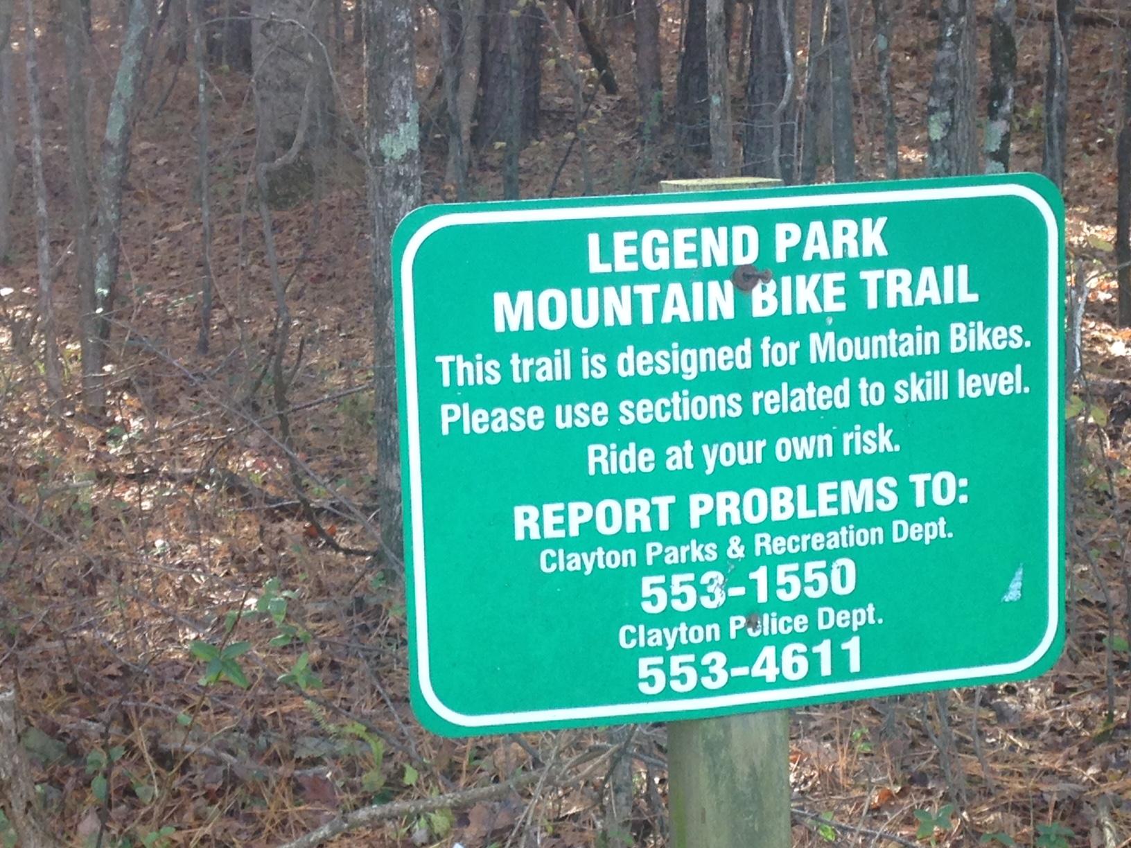 Green sign at Legend Park Mountain Bike Trail indicating that the trail is designated for mountain bikes, advising users to ride according to their skill level and to ride at their own risk. The sign provides contact information for reporting problems to the Clayton Parks & Recreation Department and the Clayton Police Department. The background features trees and underbrush typical of a wooded area. Legend Park mountain bike trail.