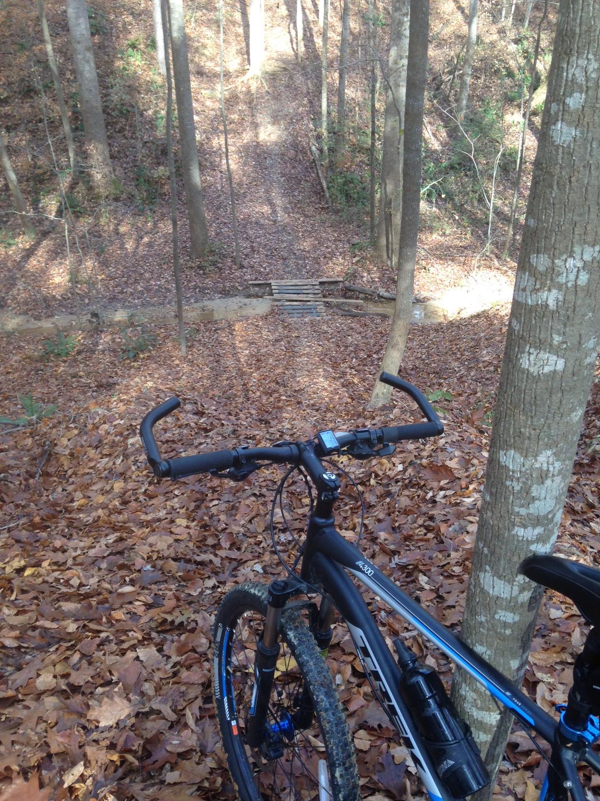 A mountain bike positioned near a dirt trail surrounded by trees, covered in fallen leaves. In the background, a wooden bridge crosses a small stream, leading deeper into the wooded area. The scene captures a serene and adventurous atmosphere for outdoor cycling. Legend Park mountain bike trail.