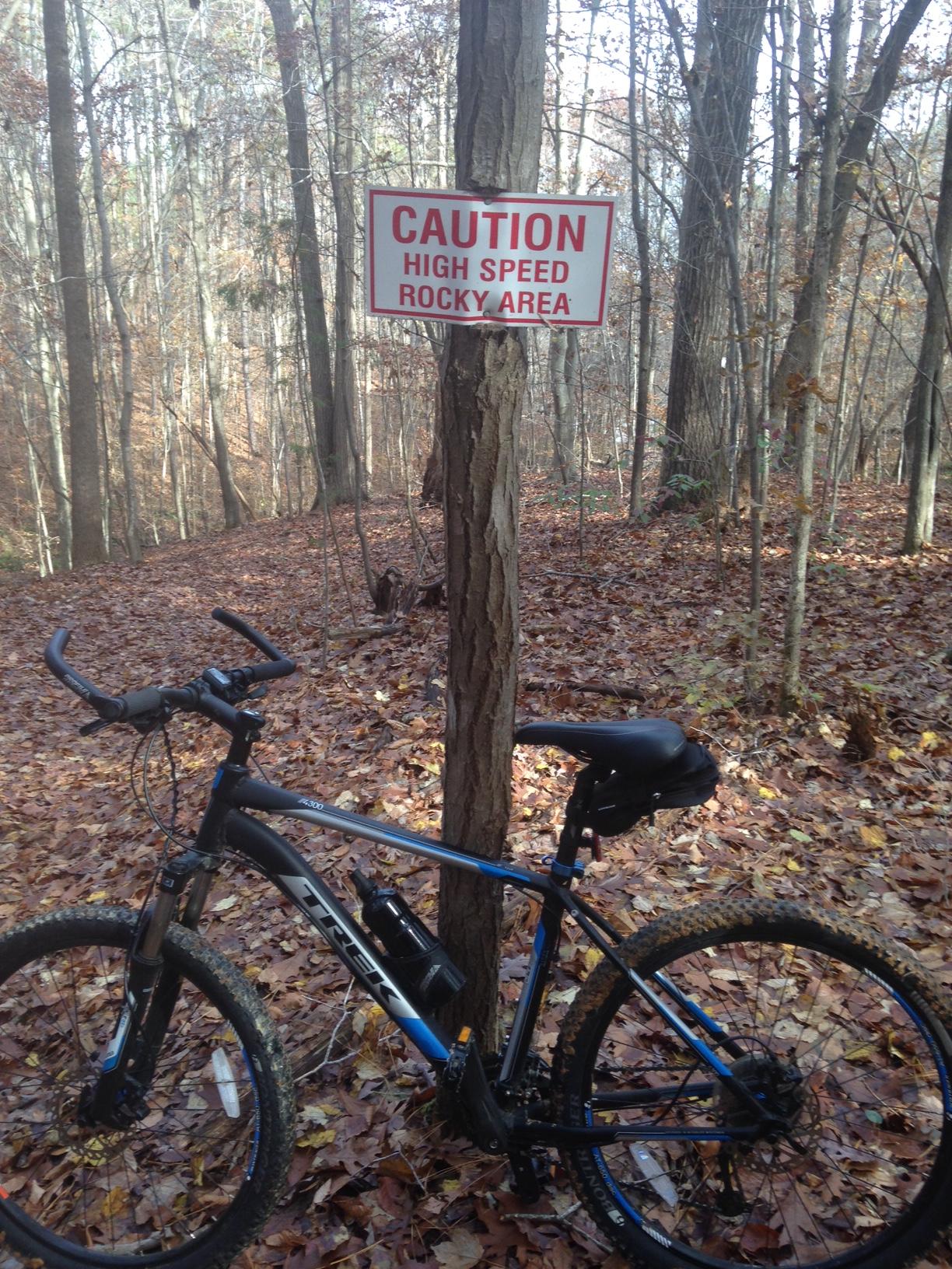 A mountain bike parked near a caution sign that reads "CAUTION HIGH SPEED ROCKY AREA," situated in a wooded area with fallen leaves on the ground and trees in the background. Legend Park mountain bike trail.