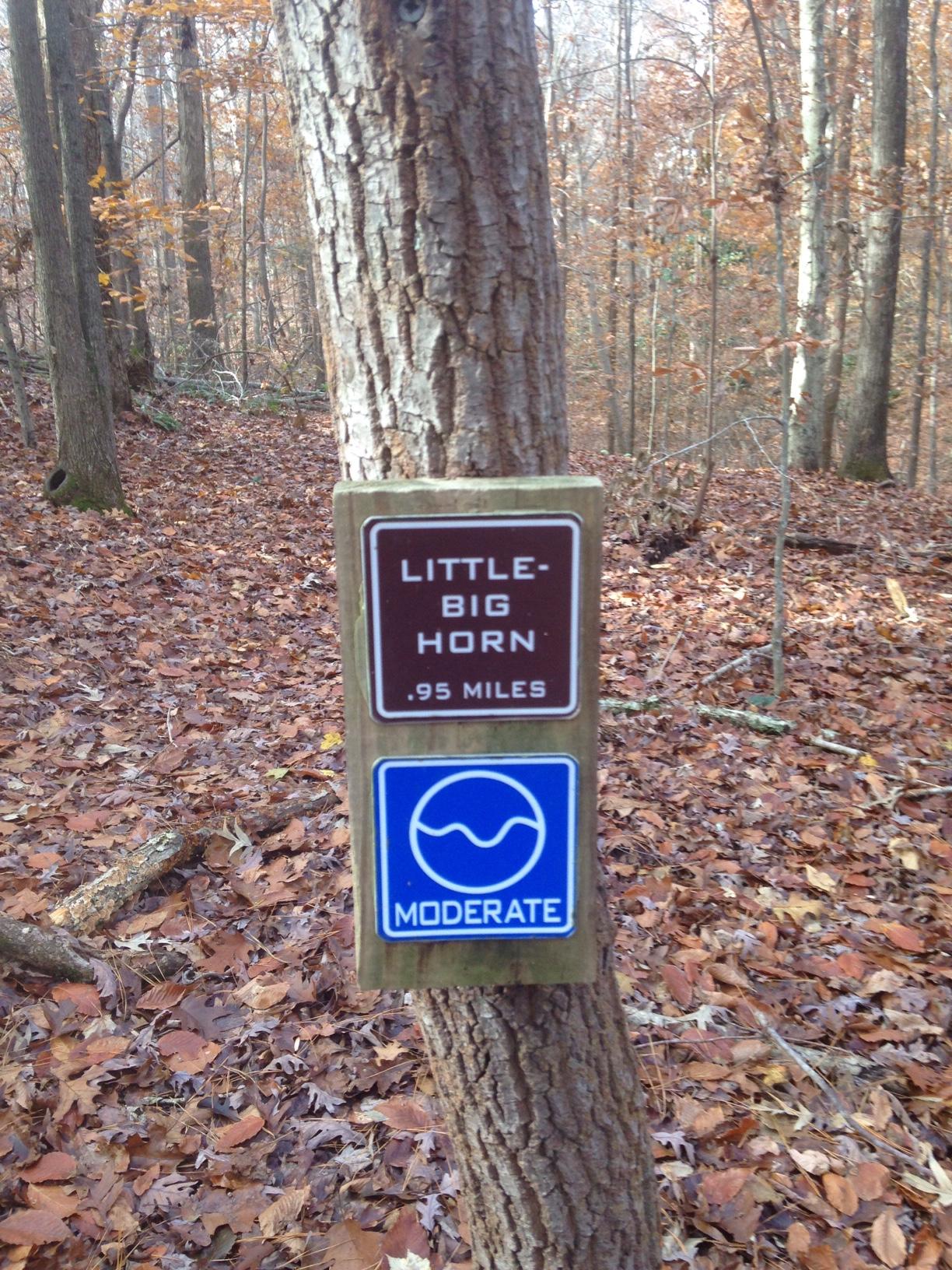 Signpost for "Little Big Horn" trail, indicating a distance of 0.95 miles and a moderate difficulty level, located in a wooded area with autumn leaves on the ground. Legend Park mountain bike trail.