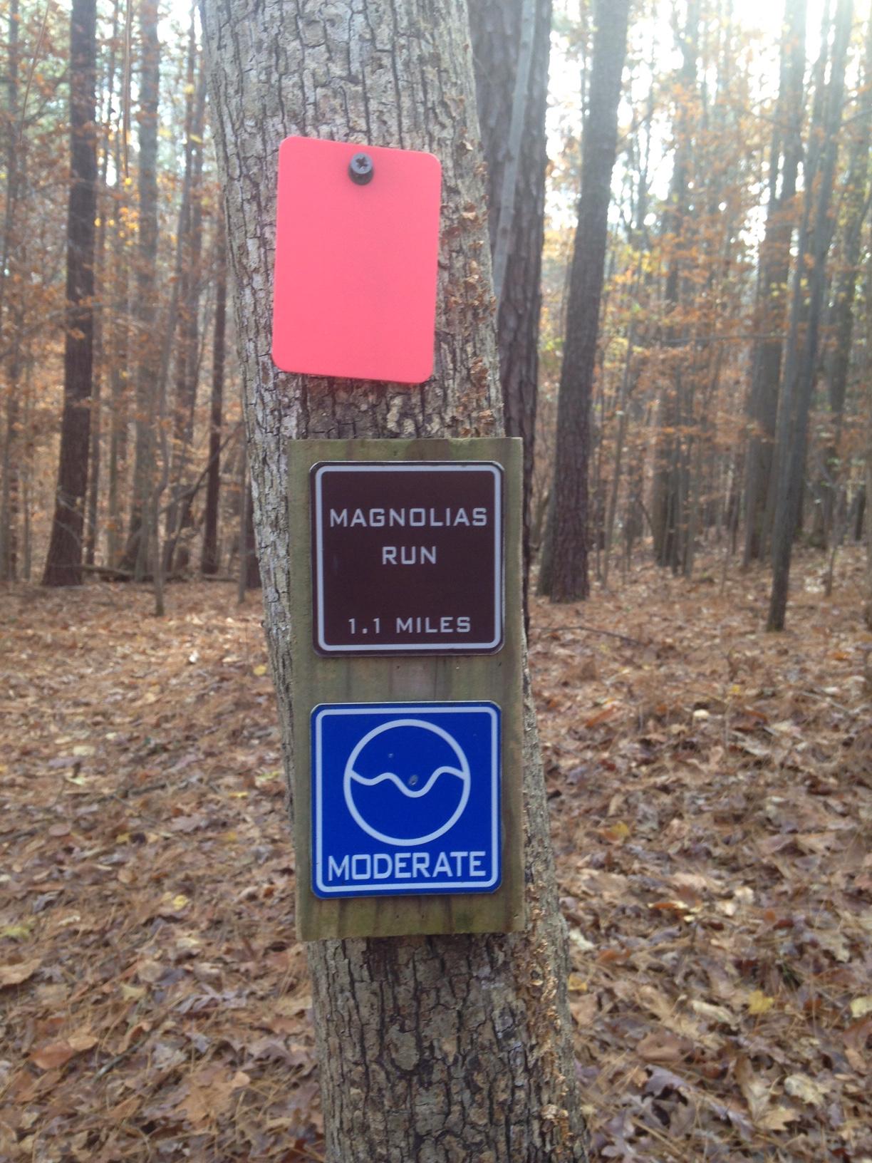 Signpost indicating "Magnolias Run," a trail that is 1.1 miles long and marked as moderate difficulty, attached to a tree in a forested area with fallen leaves. A small red tag is also visible above the sign. Legend Park mountain bike trail.
