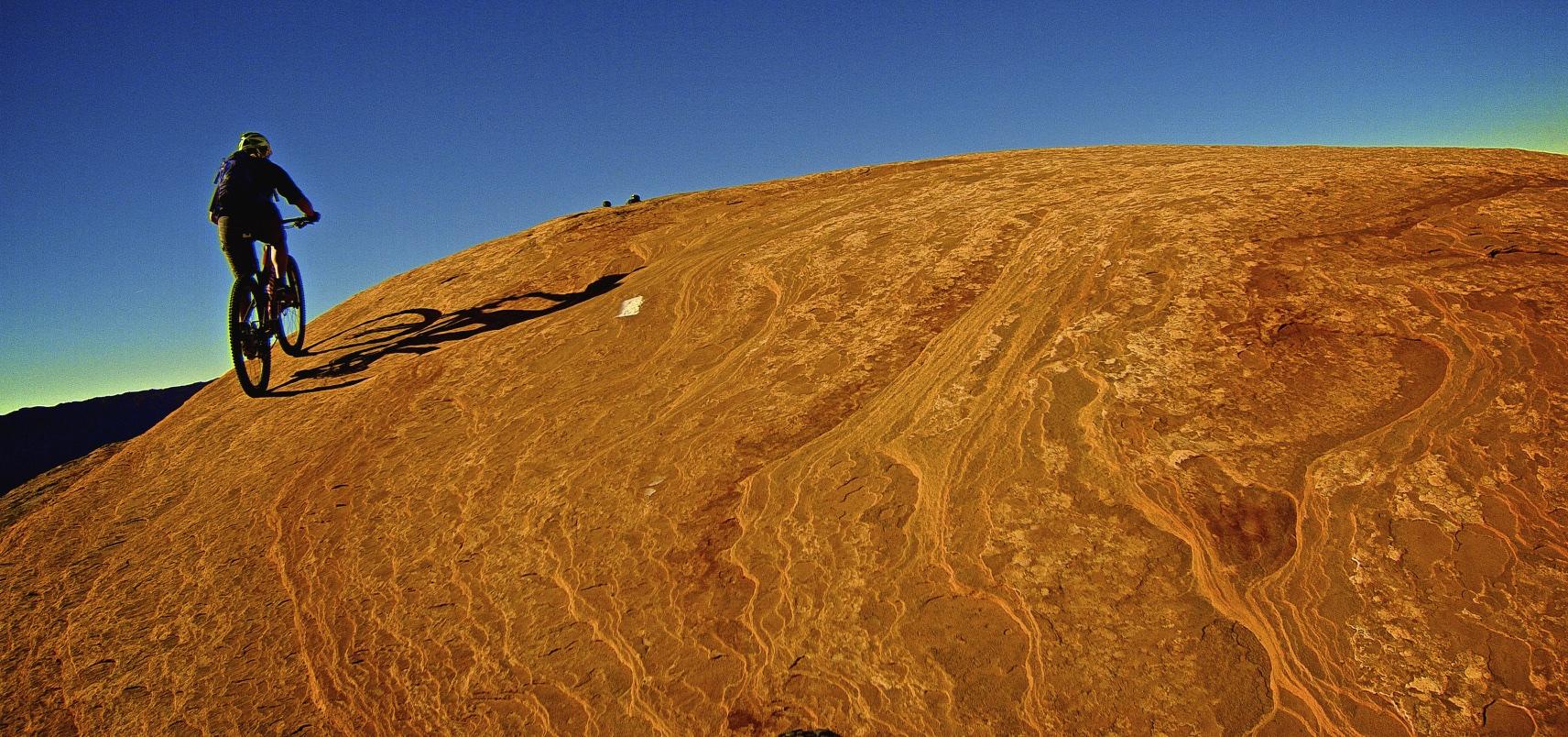 A mountain biker riding up a steep, rocky landscape under a clear blue sky. The terrain features distinct patterning and textures, illustrating the natural beauty of the setting. Shadows cast by the biker and bicycle enhance the visual interest of the scene. Slickrock mountain bike trail.