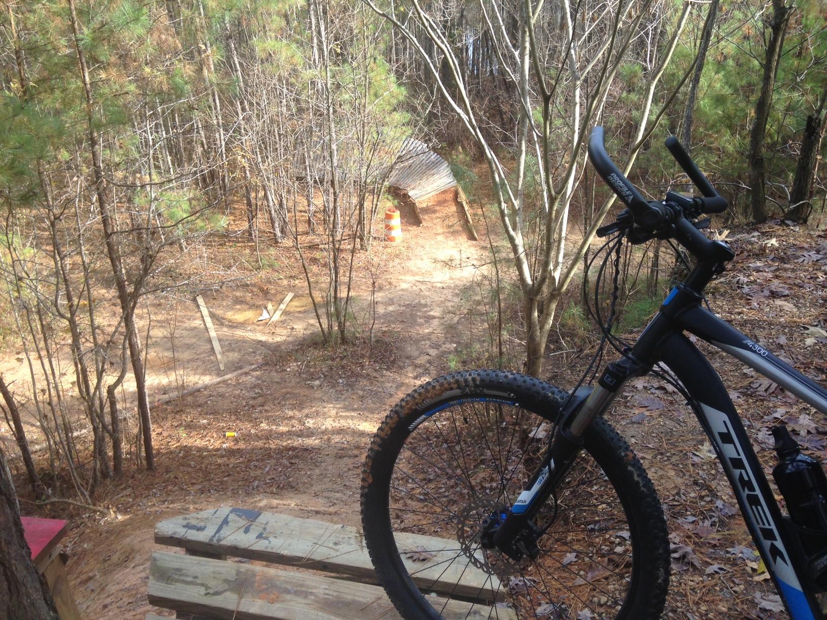 alt tag: A mountain bike is positioned on a wooden platform overlooking a dirt trail winding through a forest. The setting features sparse trees with few leaves, and a construction cone is visible in the background near a structure, indicating potential trail work or adjustments. Legend Park mountain bike trail.