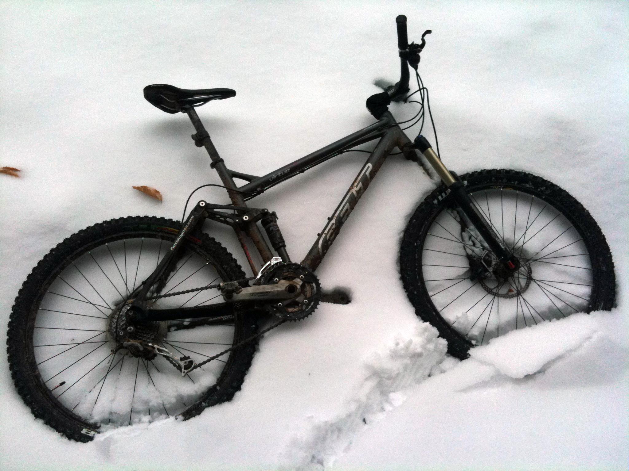 Felt Virtue Two: A mountain bike partially buried in fresh snow, with a few fallen leaves visible nearby. The bike features a sturdy frame, knobby tires, and a visible gear system, highlighting its use for off-road cycling. The surrounding snow is undisturbed except for a small area near the bike where the snow has been disturbed.