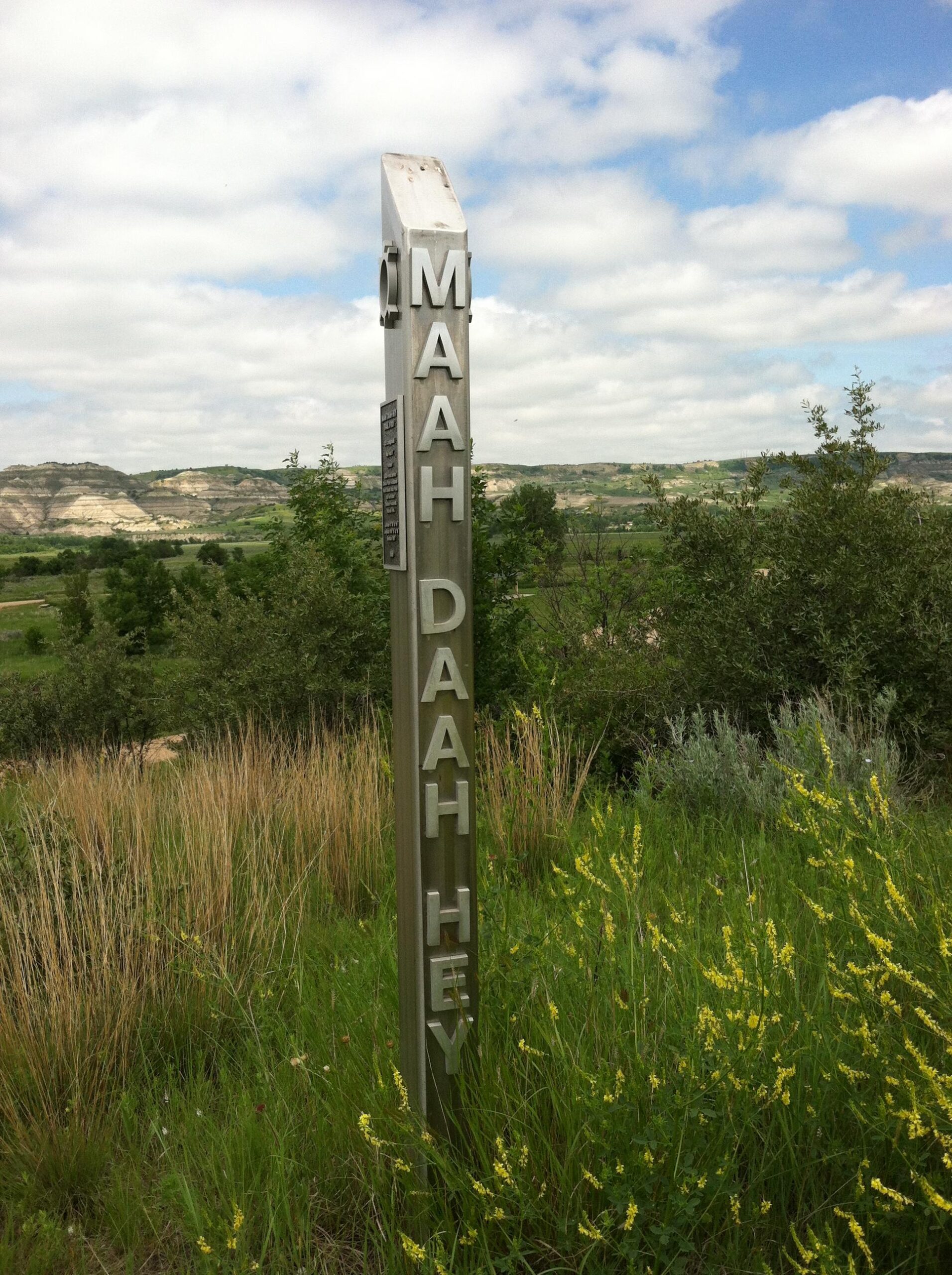 A tall brown trail marker post with the words "MAAH DAH HEY" engraved on it, standing in a grassy area with wildflowers. In the background, rolling hills under a partly cloudy sky are visible, indicating a scenic outdoor environment. Maah Daah Hey mountain bike trail.