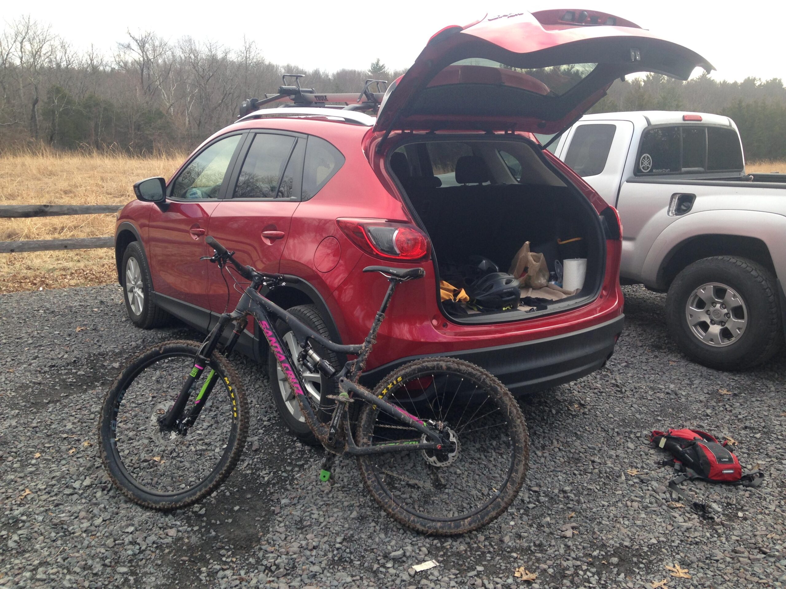 Santa Cruz Bronson: A red SUV parked on gravel with its trunk open, revealing gear and equipment inside. A mountain bike with muddy tires is positioned next to the SUV, while a second vehicle, a white truck, is visible in the background. The scene is set in a natural area with trees and dry grass.