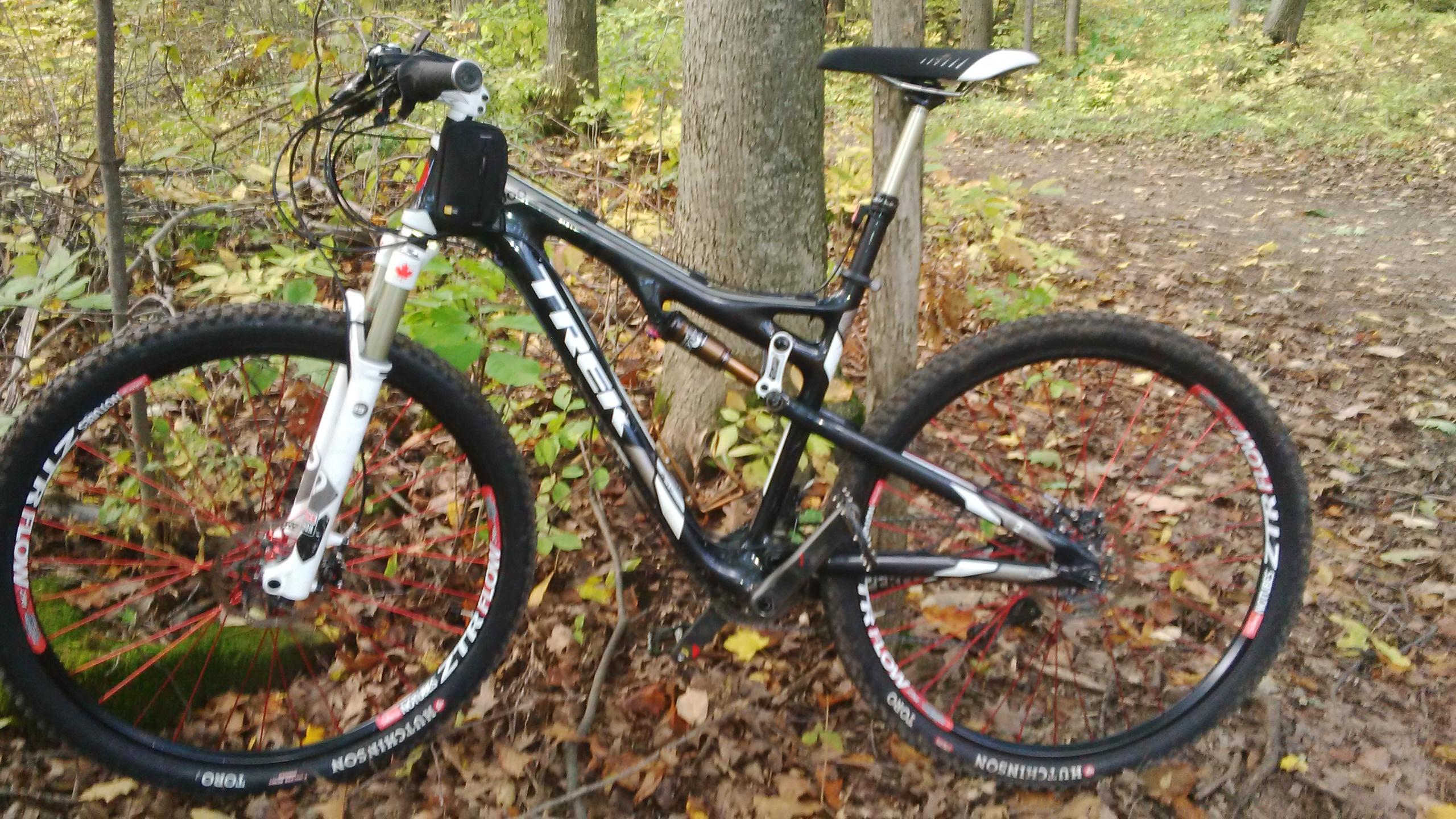 Trek Superfly Full Suspension: A black and white mountain bike with red accents parked beside a tree in a wooded area. The ground is covered with fallen leaves, and there is a dirt path visible in the background, suggesting a natural outdoor setting suitable for biking.