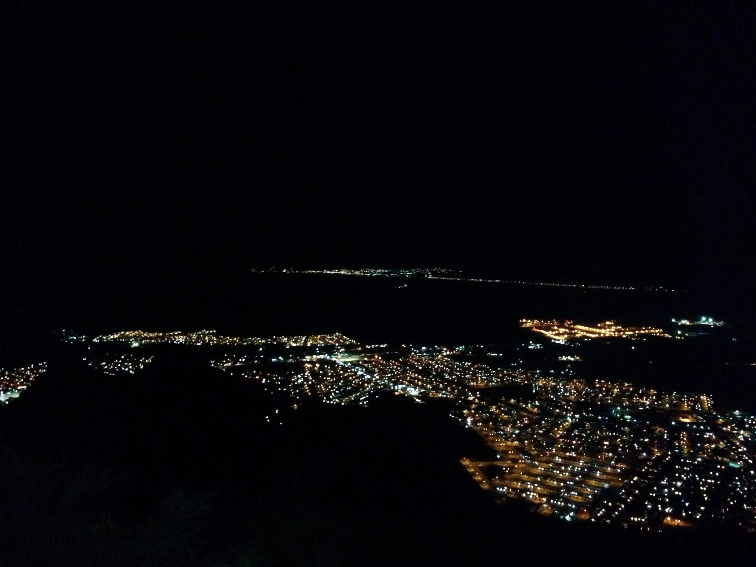 A nighttime view of a city illuminated by streetlights and buildings, with a dark sky above. The lights create a sparkling effect across the landscape, highlighting the layout of the streets and structures. In the distance, more lights can be seen along the shoreline, adding a sense of depth to the scene. Microondas mountain bike trail.