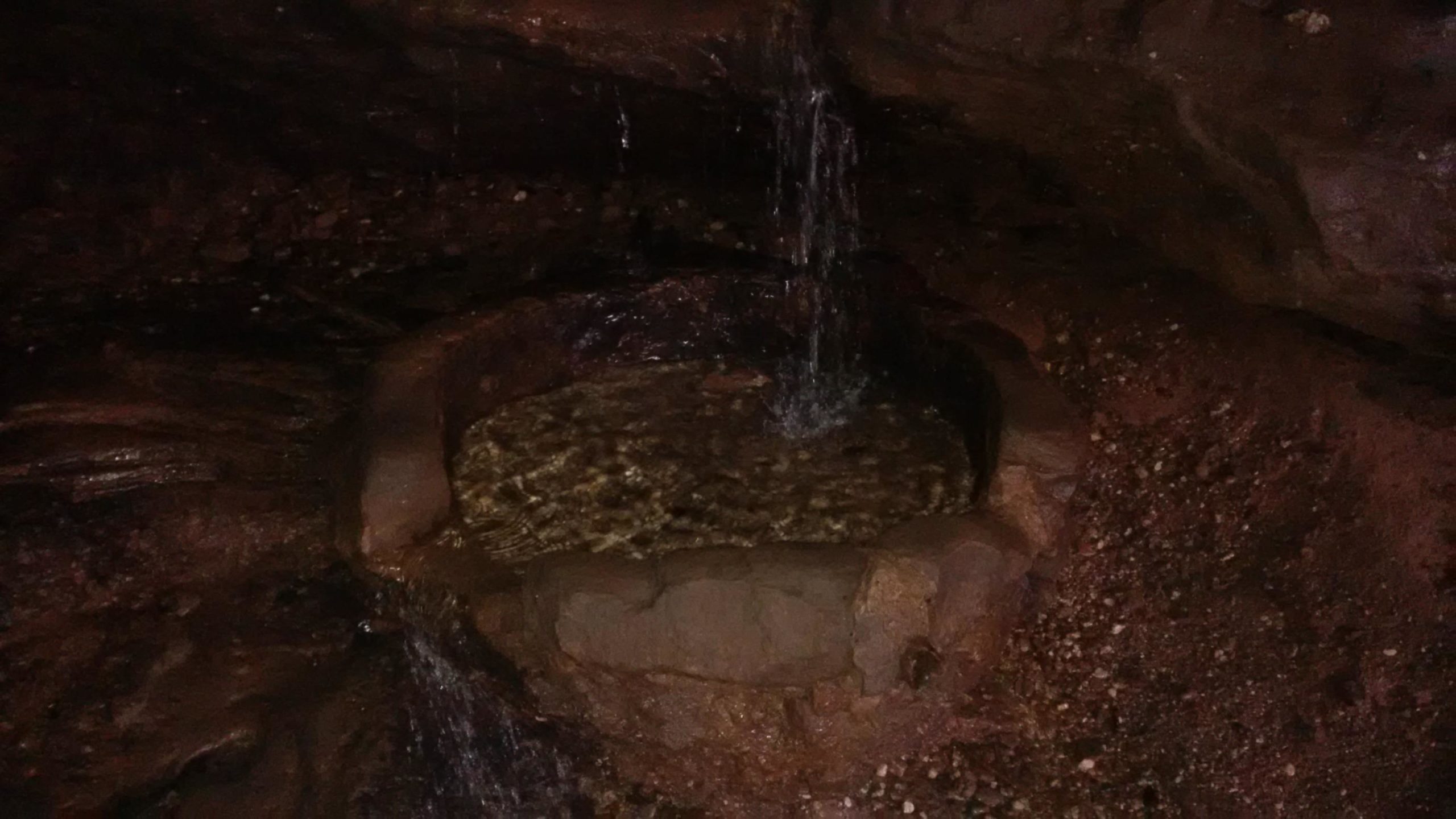 A small, circular stone basin partially filled with clear water, nestled in a rocky cave setting. Water gently flows into the basin from above, creating a serene atmosphere. The surrounding area is dimly lit, highlighting the earthy tones of the rocks and gravel. Rodalben mountain bike trail.