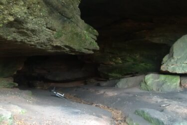 Alt text: A natural cave interior featuring stone walls with moss and foliage. The ground is uneven, covered with dirt and leaves, and a bicycle lies on its side in the foreground. Soft lighting gives the cave a shadowy appearance. Rodalben mountain bike trail.