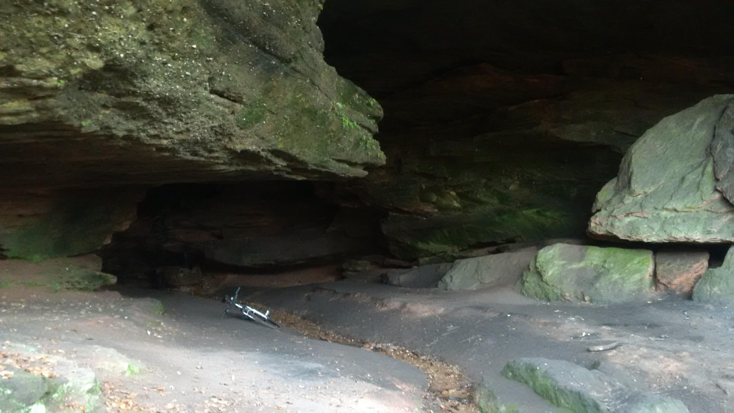 Alt text: A natural cave interior featuring stone walls with moss and foliage. The ground is uneven, covered with dirt and leaves, and a bicycle lies on its side in the foreground. Soft lighting gives the cave a shadowy appearance. Rodalben mountain bike trail.