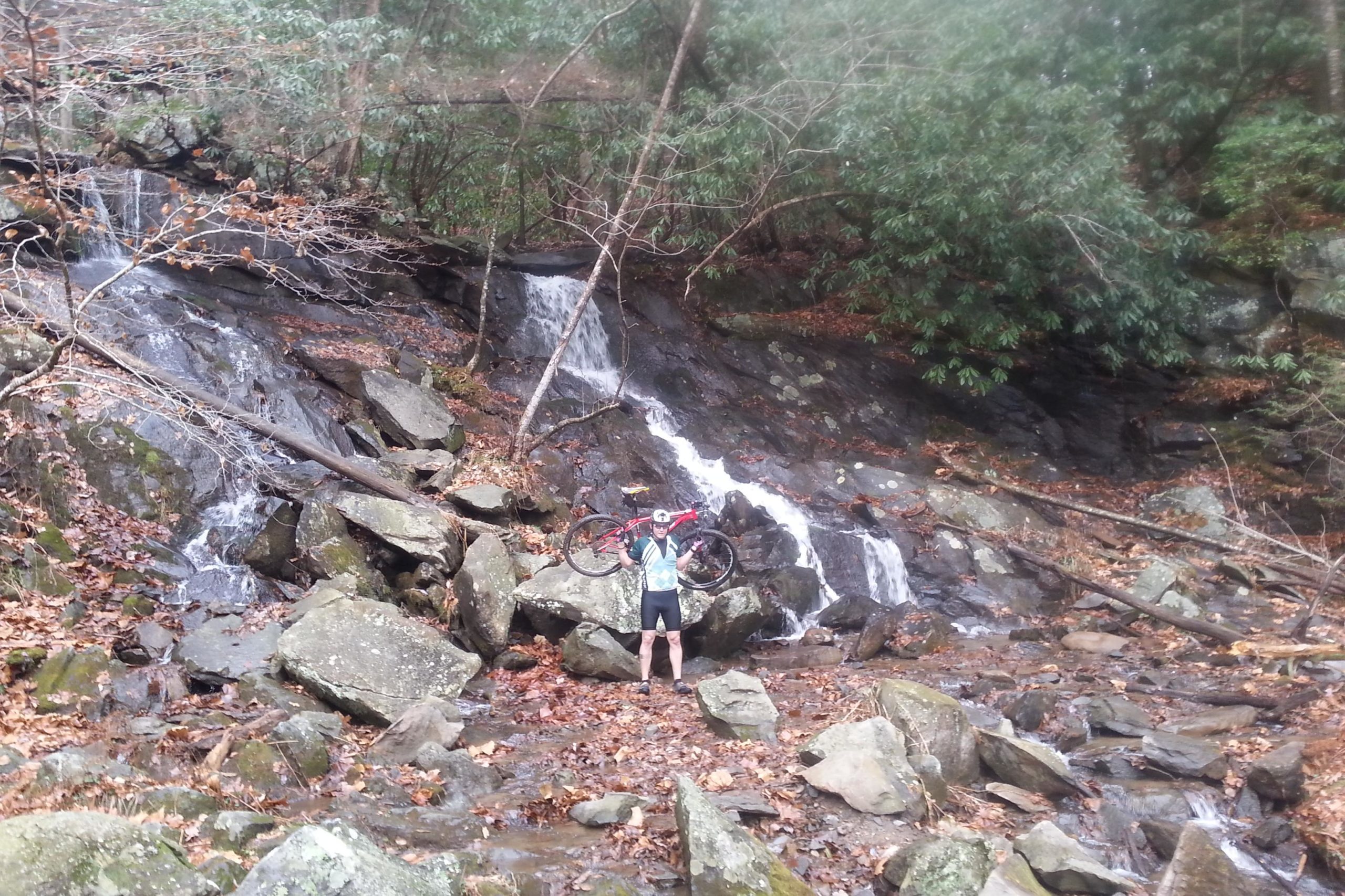 A person standing on rocky terrain near a small waterfall, holding a mountain bike above their head. The area is surrounded by trees, and the ground is covered with fallen leaves. Pinhoti Trail: P1 / Bear Creek Pinhoti mountain bike trail.