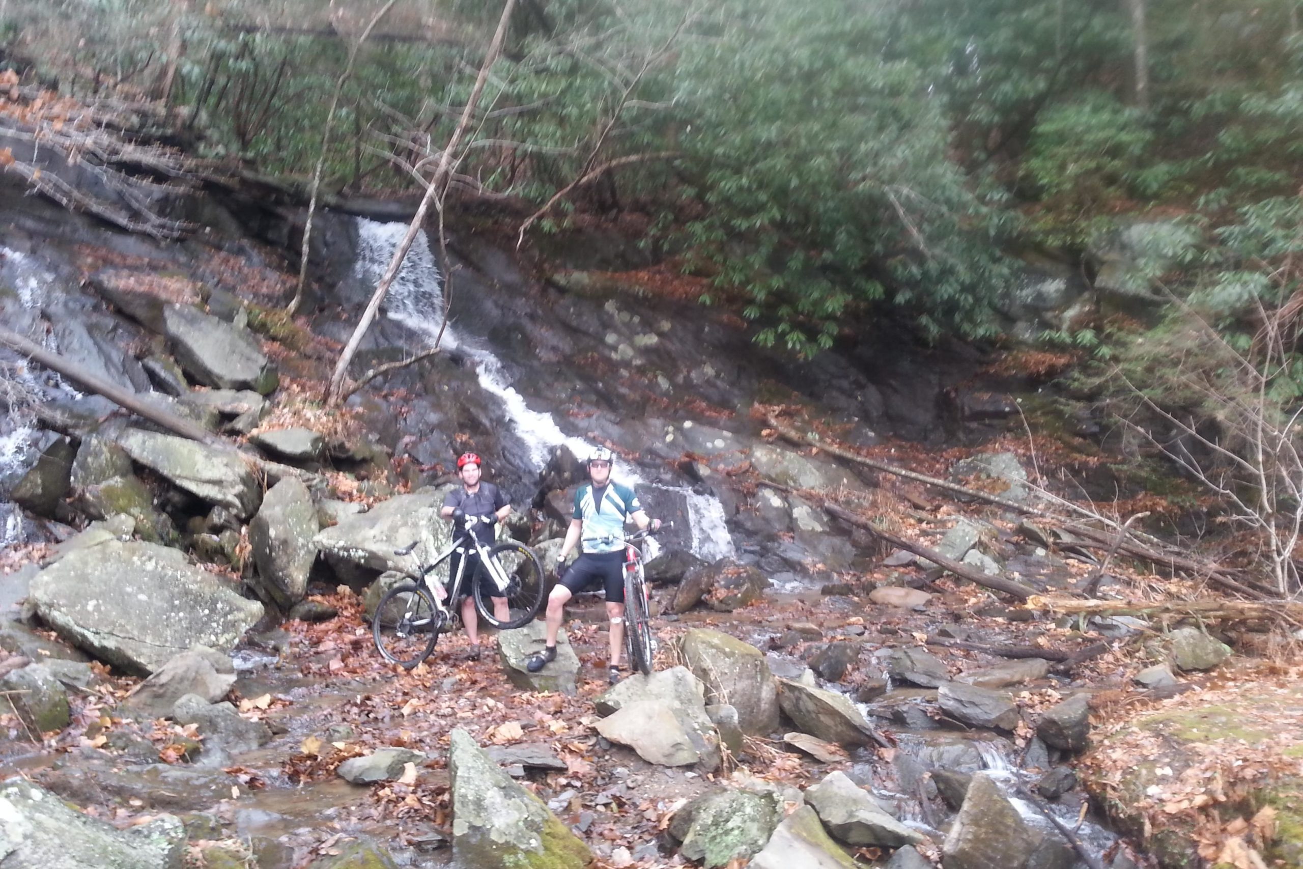 Two mountain bikers stand next to their bikes in a rocky, wooded area near a small waterfall. The ground is covered with leaves, and the surrounding trees provide a lush backdrop. Pinhoti Trail: P1 / Bear Creek Pinhoti mountain bike trail.