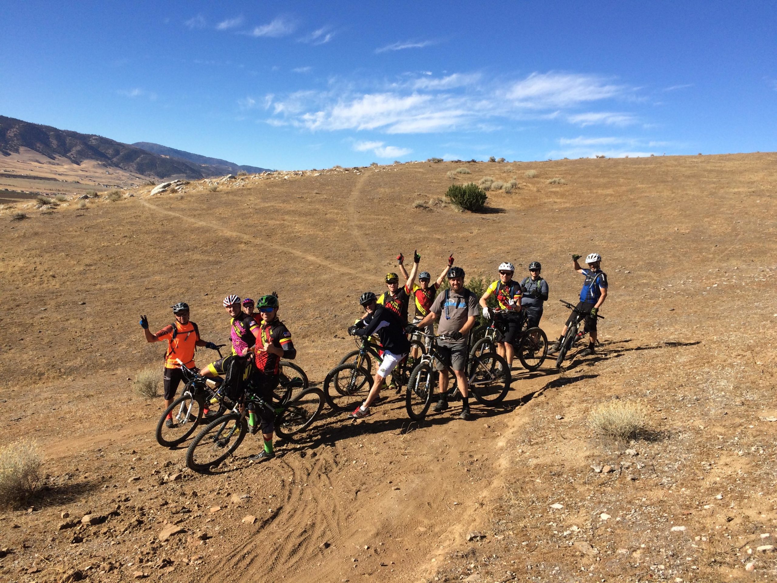 A group of mountain bikers, wearing colorful jerseys and helmets, pose for a photo on a dirt trail surrounded by dry, rocky terrain and hills under a clear blue sky. Some riders are waving or raising their hands in celebration. TMTA Lehigh trails mountain bike trail.