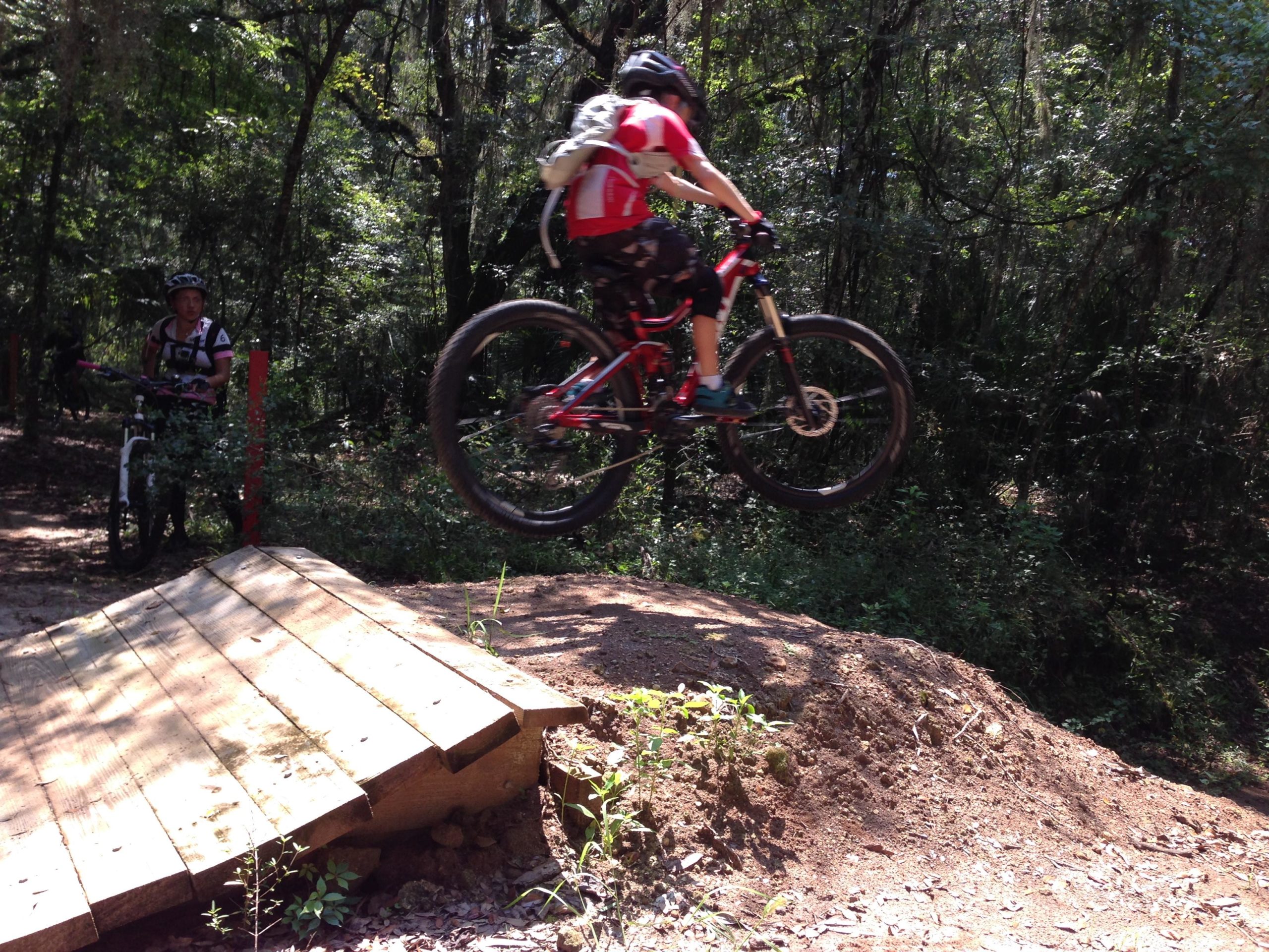 A mountain biker in a red jersey is airborne after jumping off a wooden ramp, with lush greenery surrounding the dirt trail. Another rider in the background watches. Santos mountain bike trail.