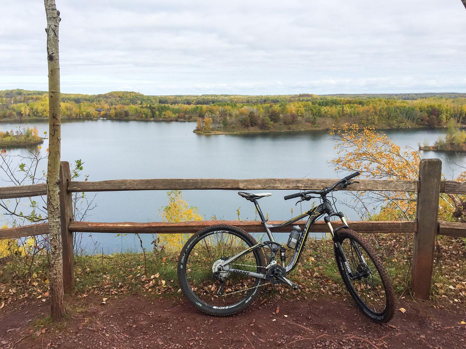 A mountain bike resting against a wooden fence with a scenic view of a calm lake surrounded by trees displaying autumn colors. The sky is overcast, enhancing the tranquil atmosphere of the landscape. Cuyuna Lakes mountain bike trail.