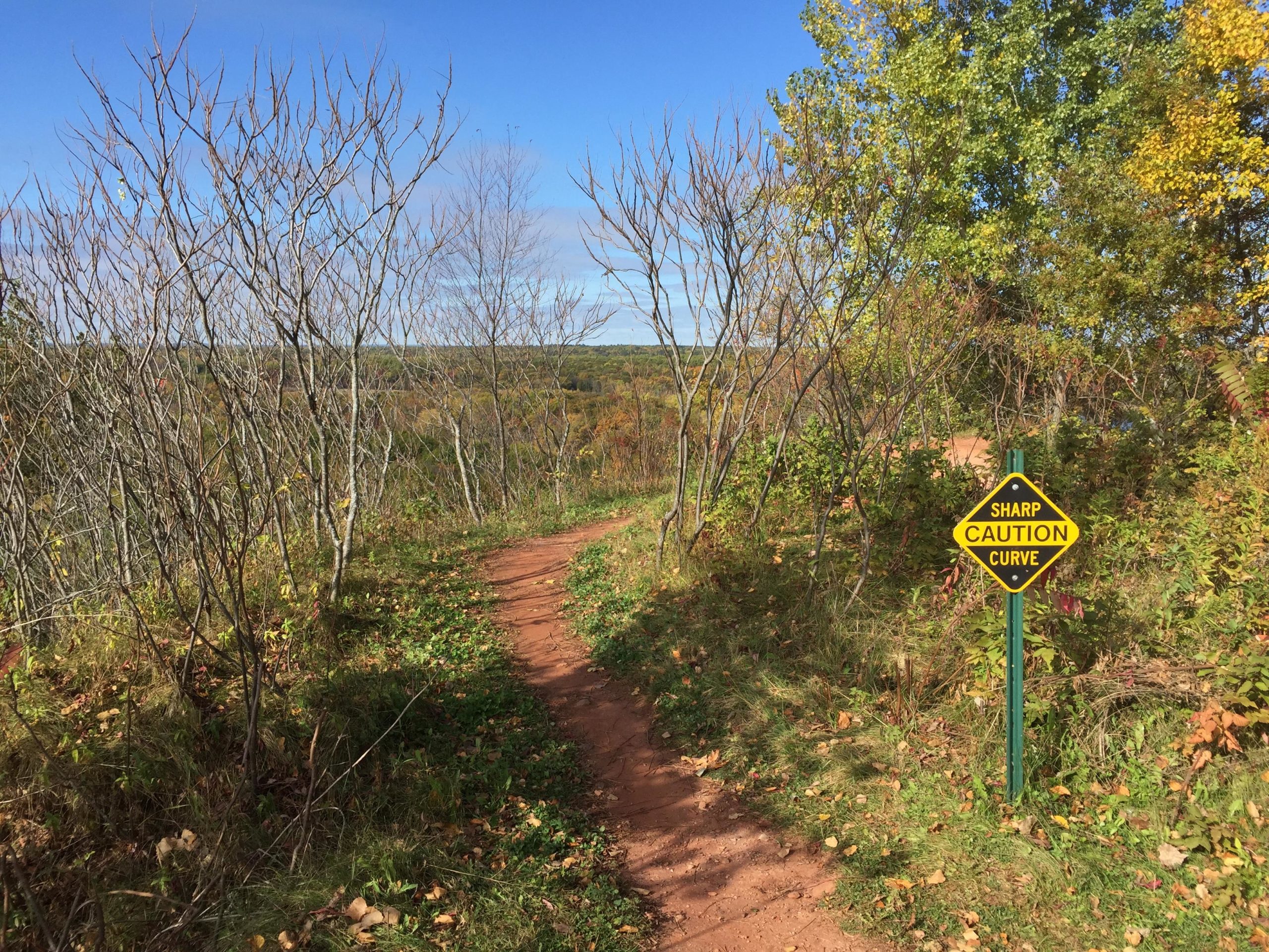 A winding dirt path surrounded by trees and shrubs, with a bright blue sky in the background. A warning sign is visible on the right, indicating a "Sharp Caution Curve." The scene captures the natural beauty of a trail, highlighting the autumn foliage and the landscape beyond. Cuyuna Lakes mountain bike trail.