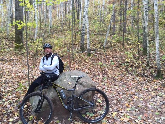 Giant Trance X 29er 2: A person wearing a cycling helmet and a jacket sits on a large rock in a forested area, with autumn leaves covering the ground. A mountain bike leans against the rock beside them, and trees with green and yellow leaves surround the scene.
