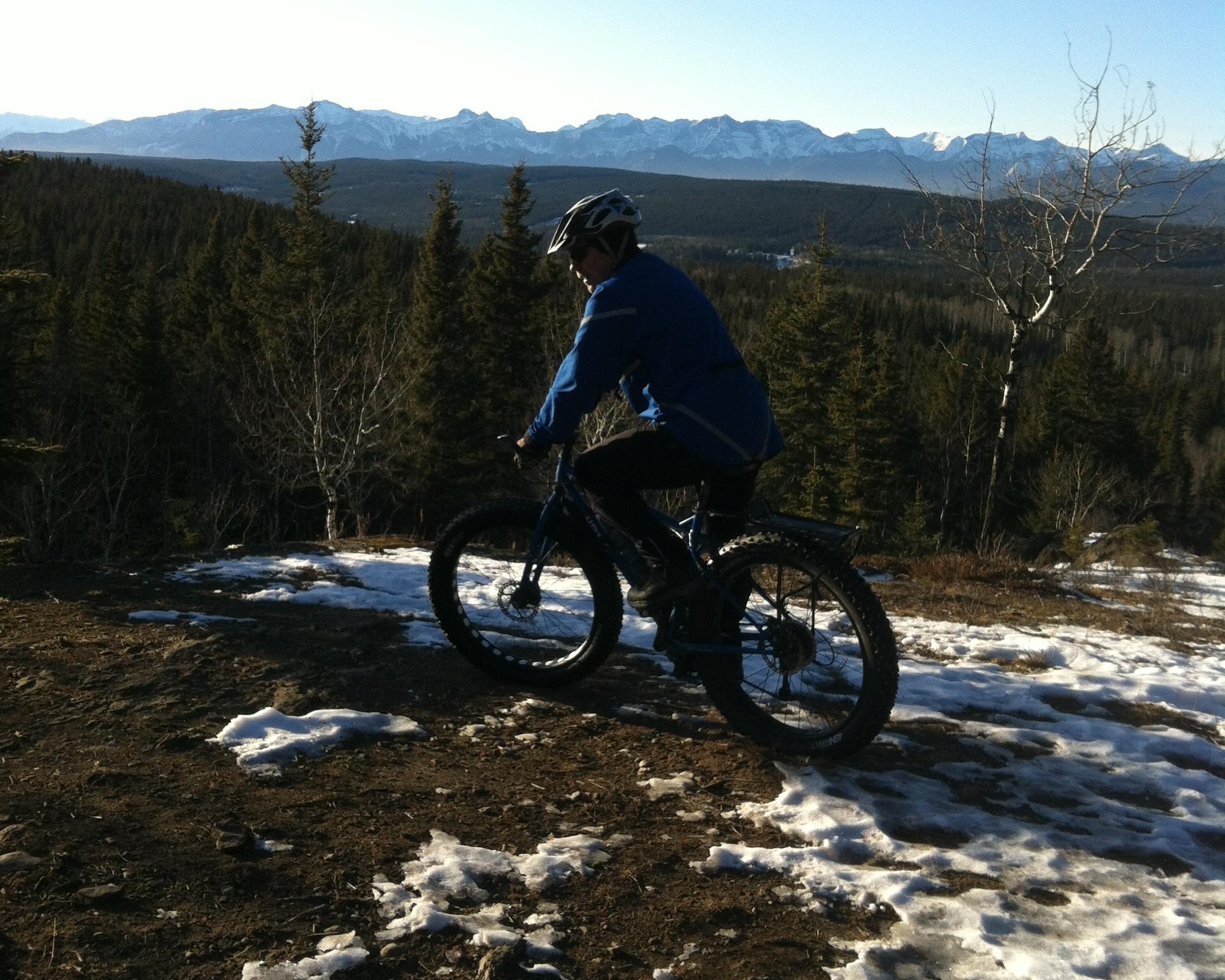 A mountain biker in a blue jacket rides on a snowy trail with a backdrop of mountains and dense forests under a clear sky. The scene captures the rugged beauty of an outdoor landscape, illustrating an active lifestyle in nature. Happy Creek Trail System mountain bike trail.