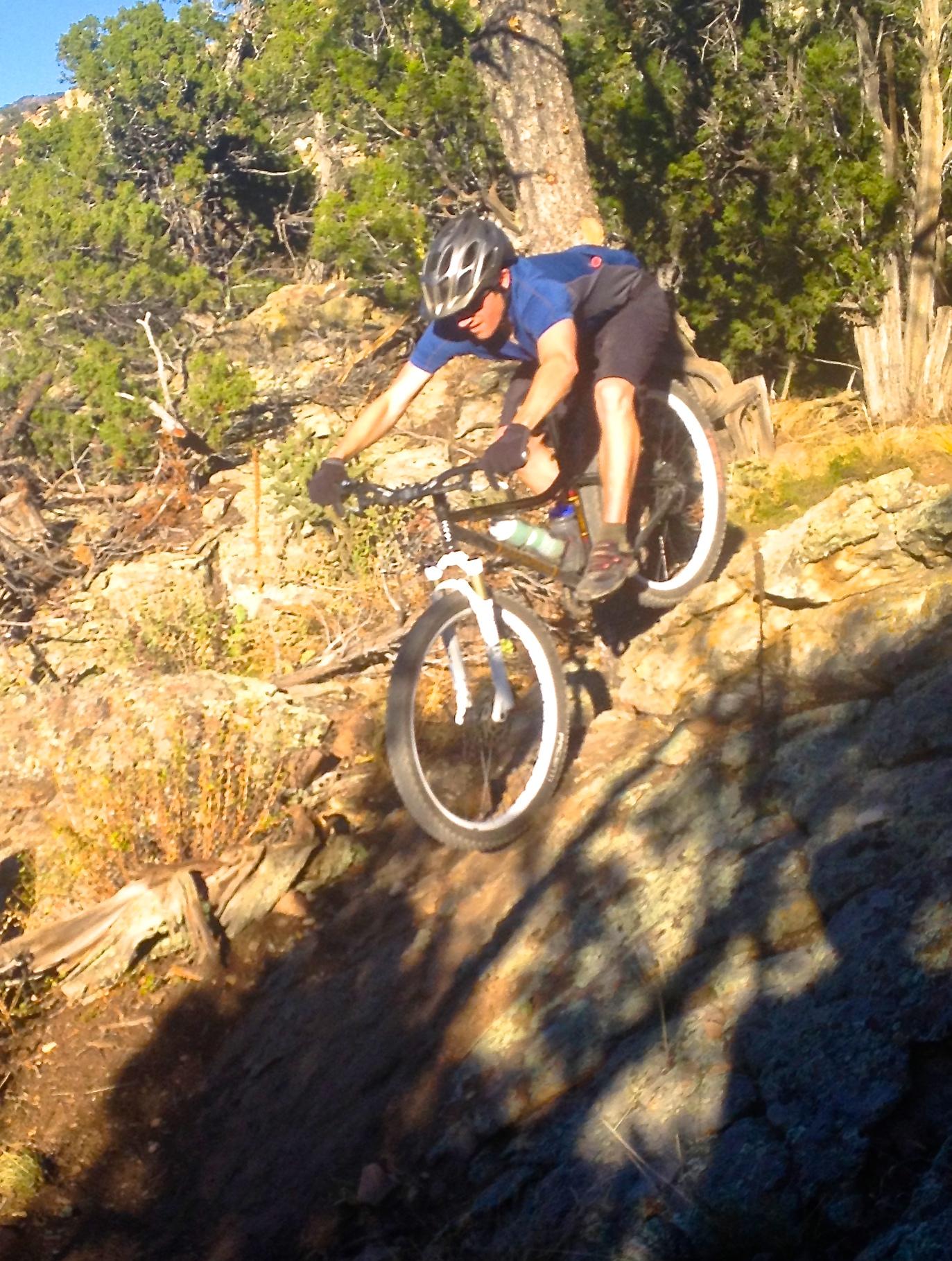 A mountain biker in a blue shirt and black shorts navigates a rocky trail, leaning forward with determination as he rides downhill on a mountain bike. Trees and natural foliage are visible in the background, highlighting the rugged outdoor environment. Oil Well Flats mountain bike trail.