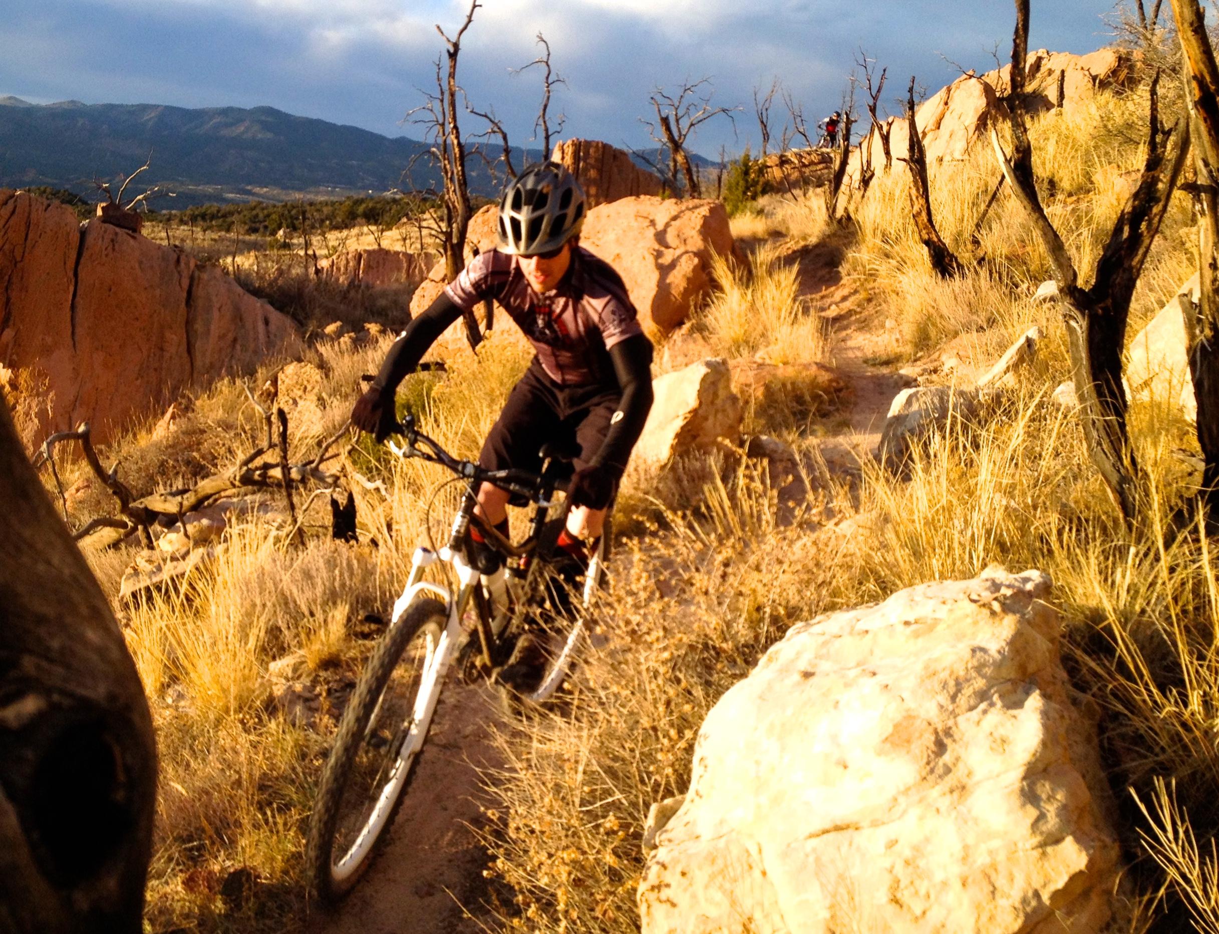 A cyclist navigating a rocky trail surrounded by dry grass and skeletal trees, with a mountainous landscape in the background under a partly cloudy sky. Oil Well Flats mountain bike trail.