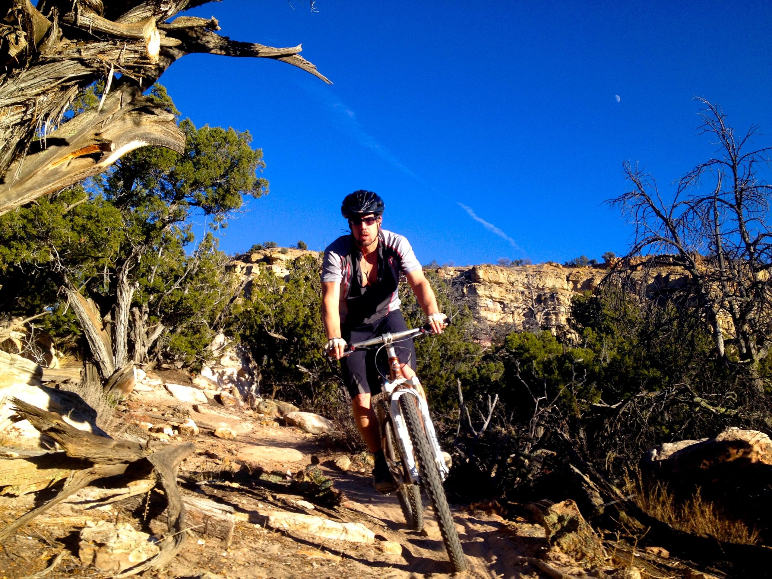 A person mountain biking on a rugged trail surrounded by trees and rocky terrain, with a clear blue sky and a visible crescent moon in the background. Oil Well Flats mountain bike trail.