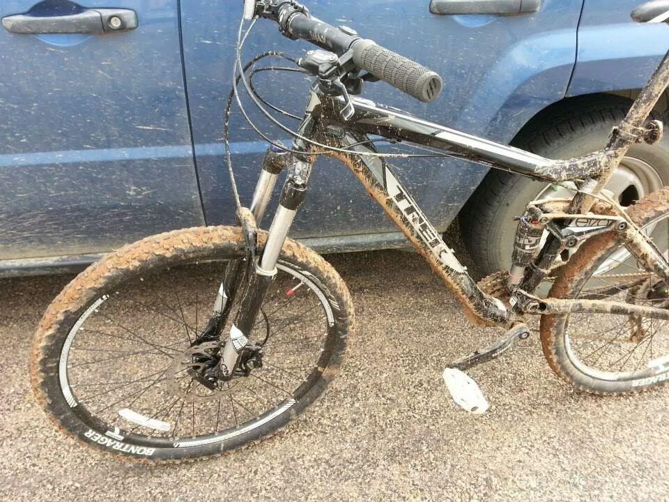 Trek Fuel EX 5: A mountain bike covered in mud leans against a blue vehicle. The bike features black and silver components, with noticeable dirt on the tires, frame, and chain. The setting appears to be an outdoor area, suggesting recent use in muddy conditions.