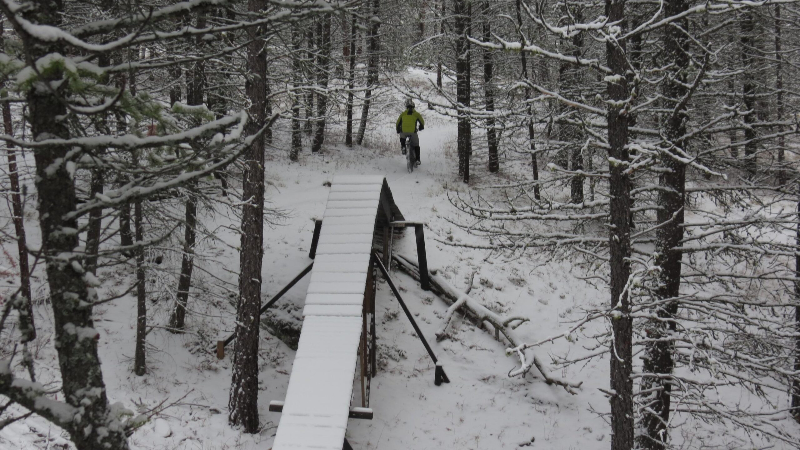 A mountain biker wearing a bright yellow jacket rides along a snowy path in a forest. A wooden bridge spans a small gap along the trail, surrounded by trees covered in fresh snow. Smith Creek mountain bike trail.