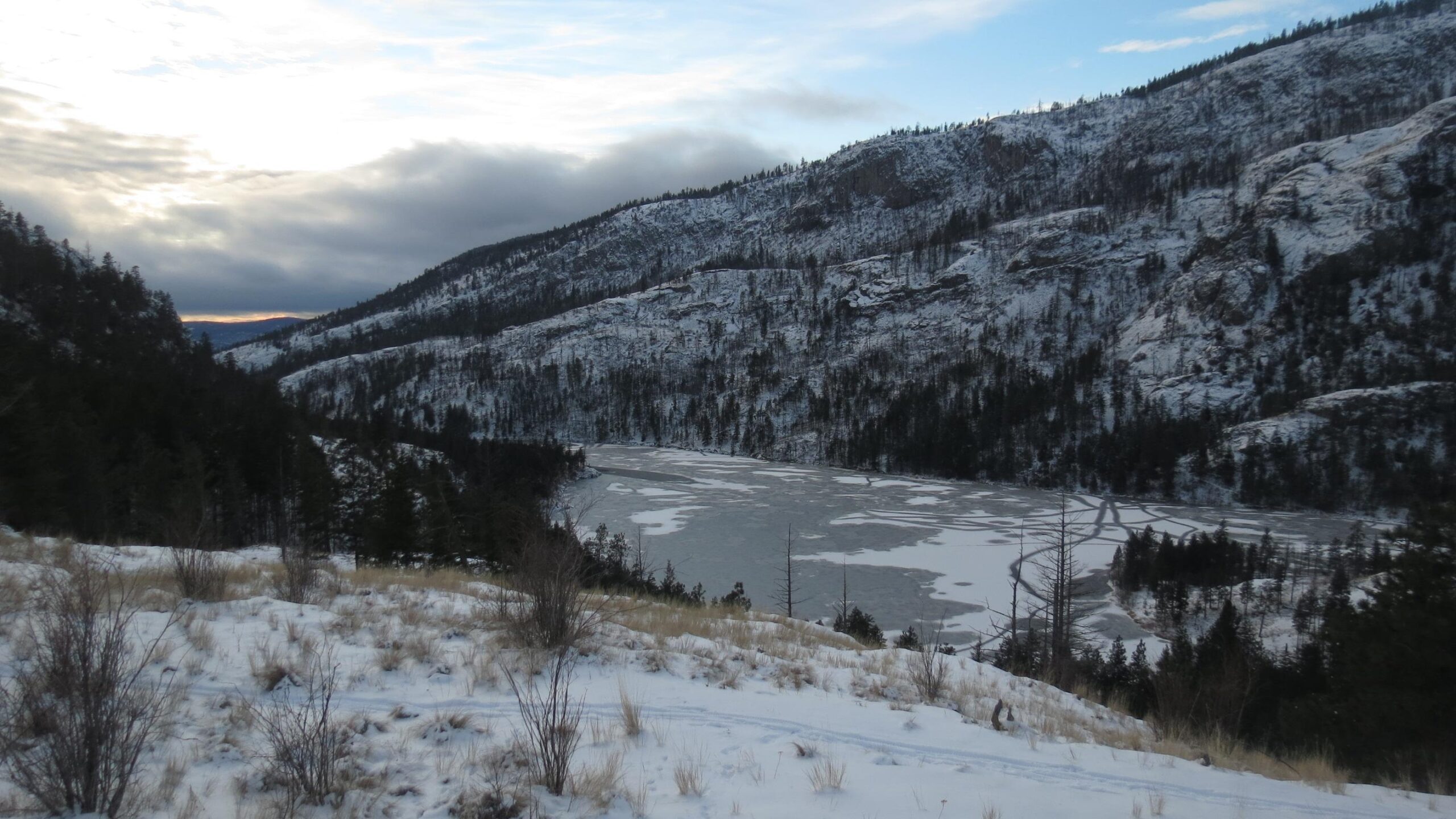 A winter landscape featuring snow-covered hills and a partially frozen lake, surrounded by evergreen trees. The sky is overcast with hints of color at the horizon, suggesting sunset. Snow blankets the ground, and the terrain is rugged, offering a serene and tranquil atmosphere. Rose Valley Resevoir mountain bike trail.