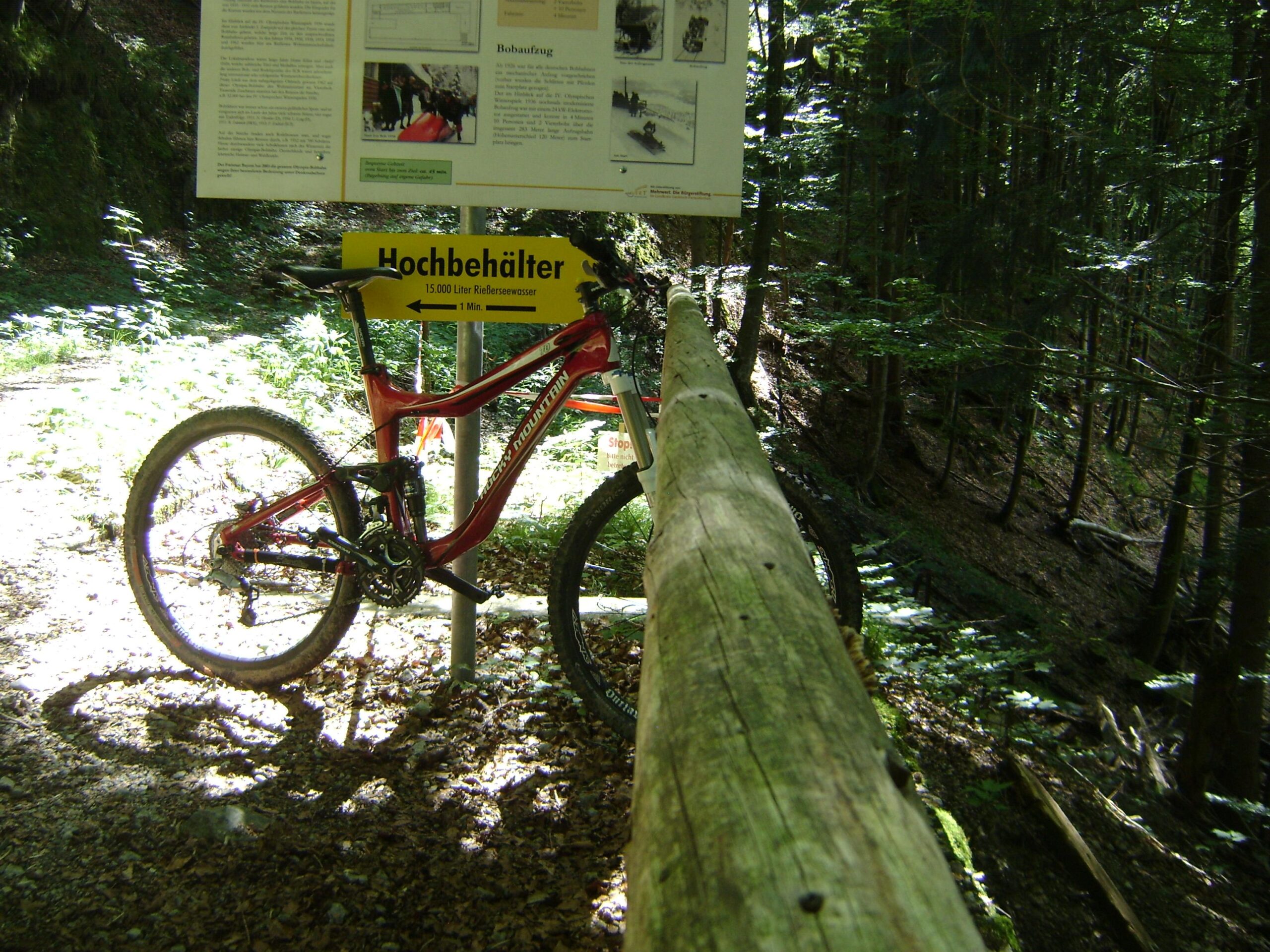 Rocky Mountain Altitude 70RSL: A red mountain bike leaning against a wooden railing in a forested area, with a sign displaying information about "Hochbehälter" and directions. Sunlight filters through the trees, casting dappled shadows on the ground.