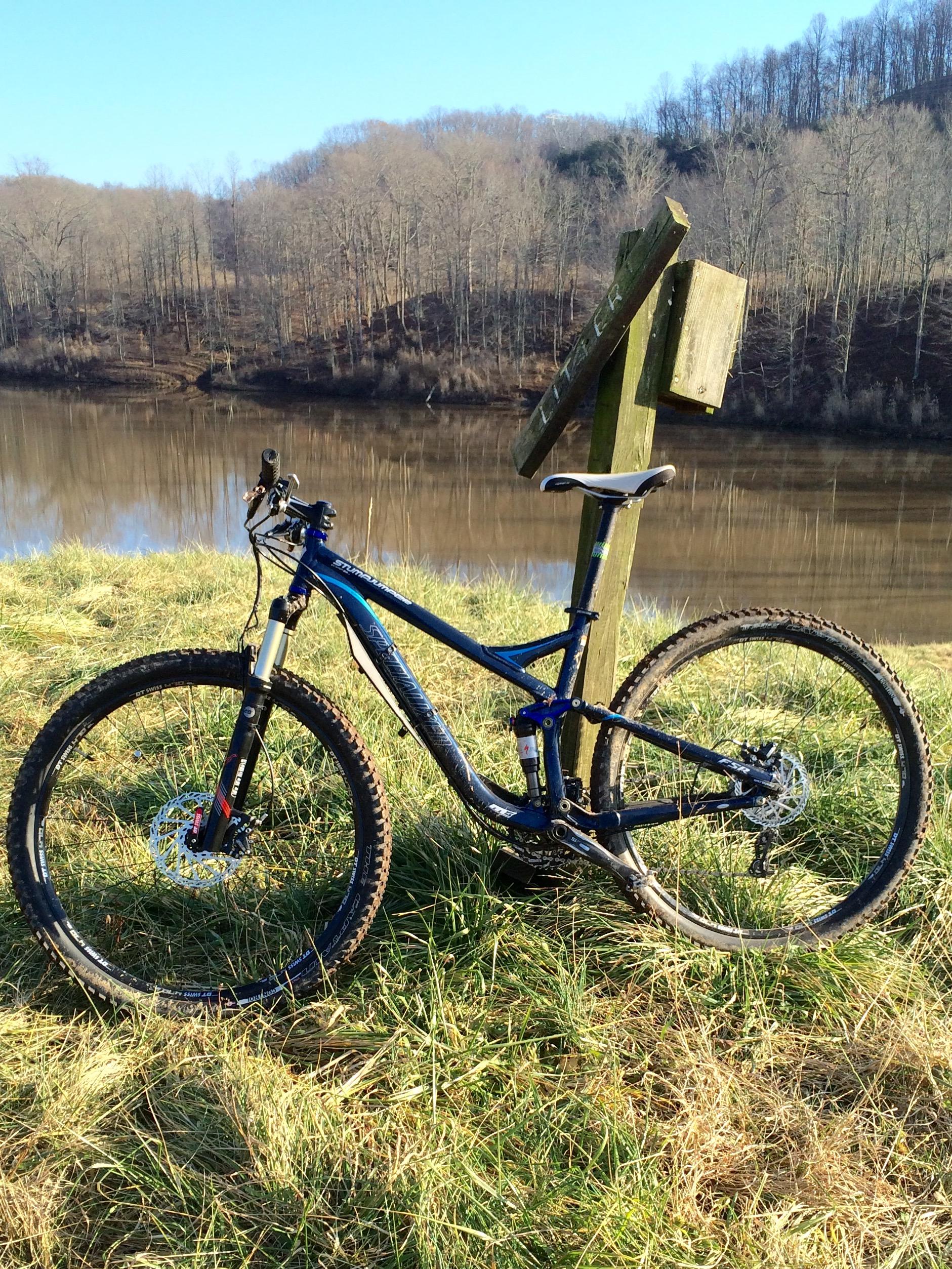 A blue mountain bike parked on grass near a river, with a wooden signpost in the background. Trees are visible in the distance, and the scene is bathed in natural sunlight, suggesting a serene outdoor environment. Mountwood mountain bike trail.