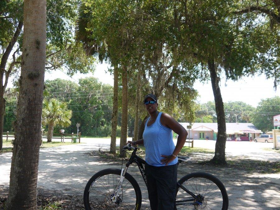 Jamis 2013 Dragon 29 Sport: A person wearing a blue tank top and sunglasses stands next to a mountain bike under a cluster of trees. The background features a dirt path and a small building, with greenery in the distance.
