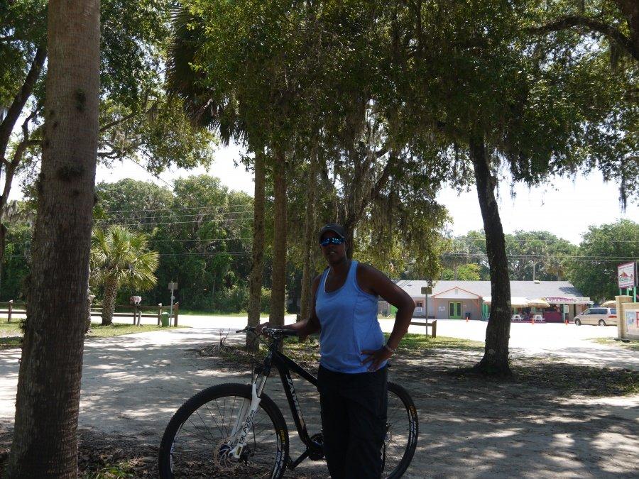Jamis 2013 Dragon 29 Sport: A person stands beside a mountain bike in a park-like setting, surrounded by trees with lush green foliage. The individual is wearing a blue tank top, black pants, and sunglasses. In the background, there are signs and hints of buildings, indicating a recreational area. The scene is bright and sunny, capturing a relaxed outdoor atmosphere.
