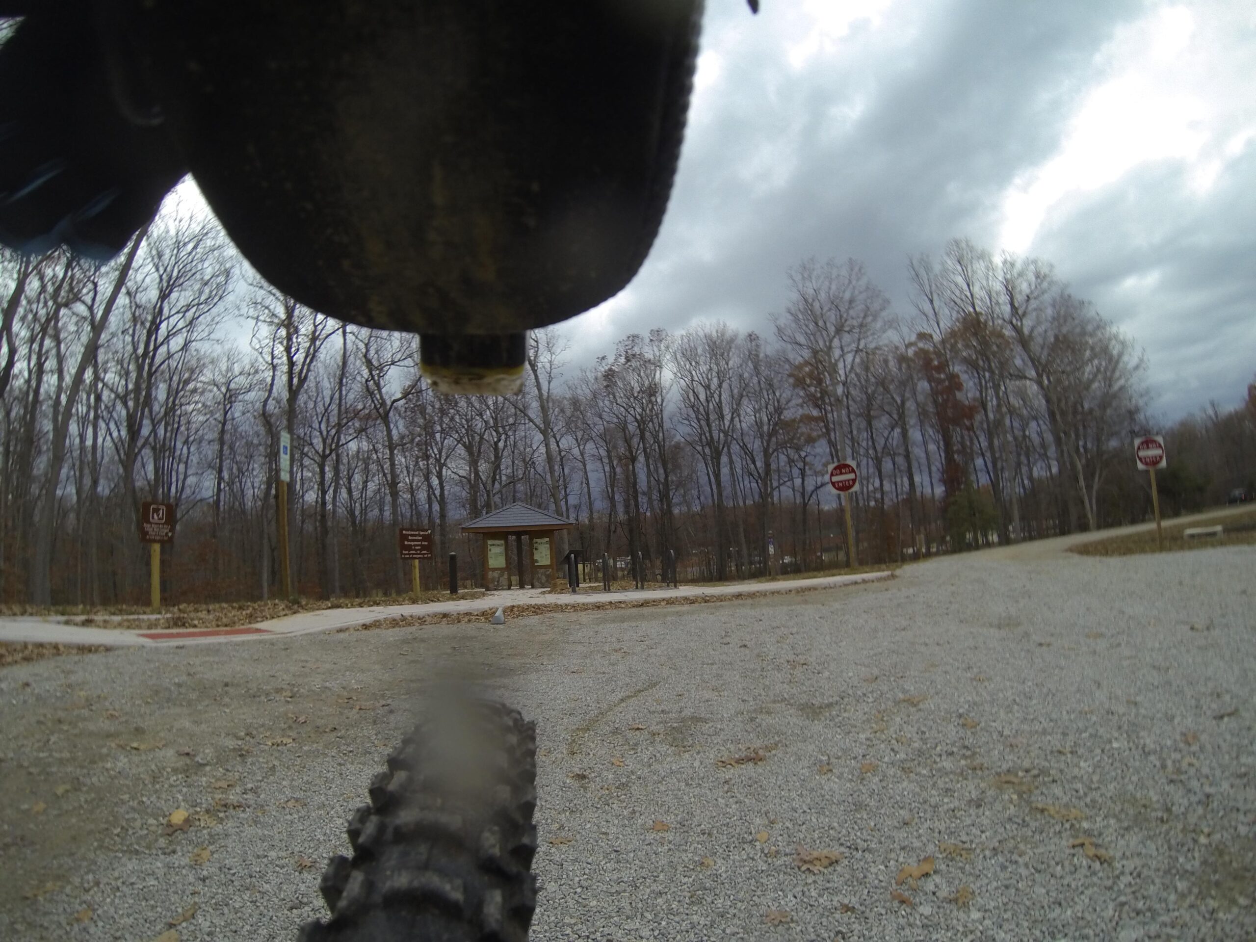 A close-up view of a mountain bike tire on gravel road with a park entrance in the background. The scene is surrounded by bare trees under a cloudy sky, with signs indicating park rules visible nearby. Meadowood mountain bike trail.
