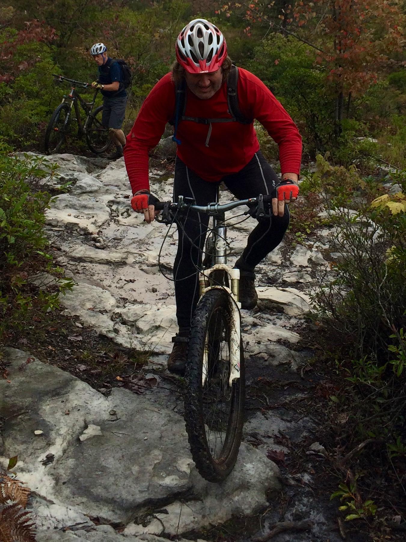 Two mountain bikers navigate a rocky trail in a wooded area. The rider in the foreground, wearing a red long-sleeve shirt and a helmet, struggles to pedal up a slippery section of the path. In the background, another cyclist, dressed in a blue shirt and gray shorts, is pushing their bike up the trail. The surroundings are lush with greenery, and the ground is wet and muddy, indicating a recent rainfall. CVI Trails mountain bike trail.